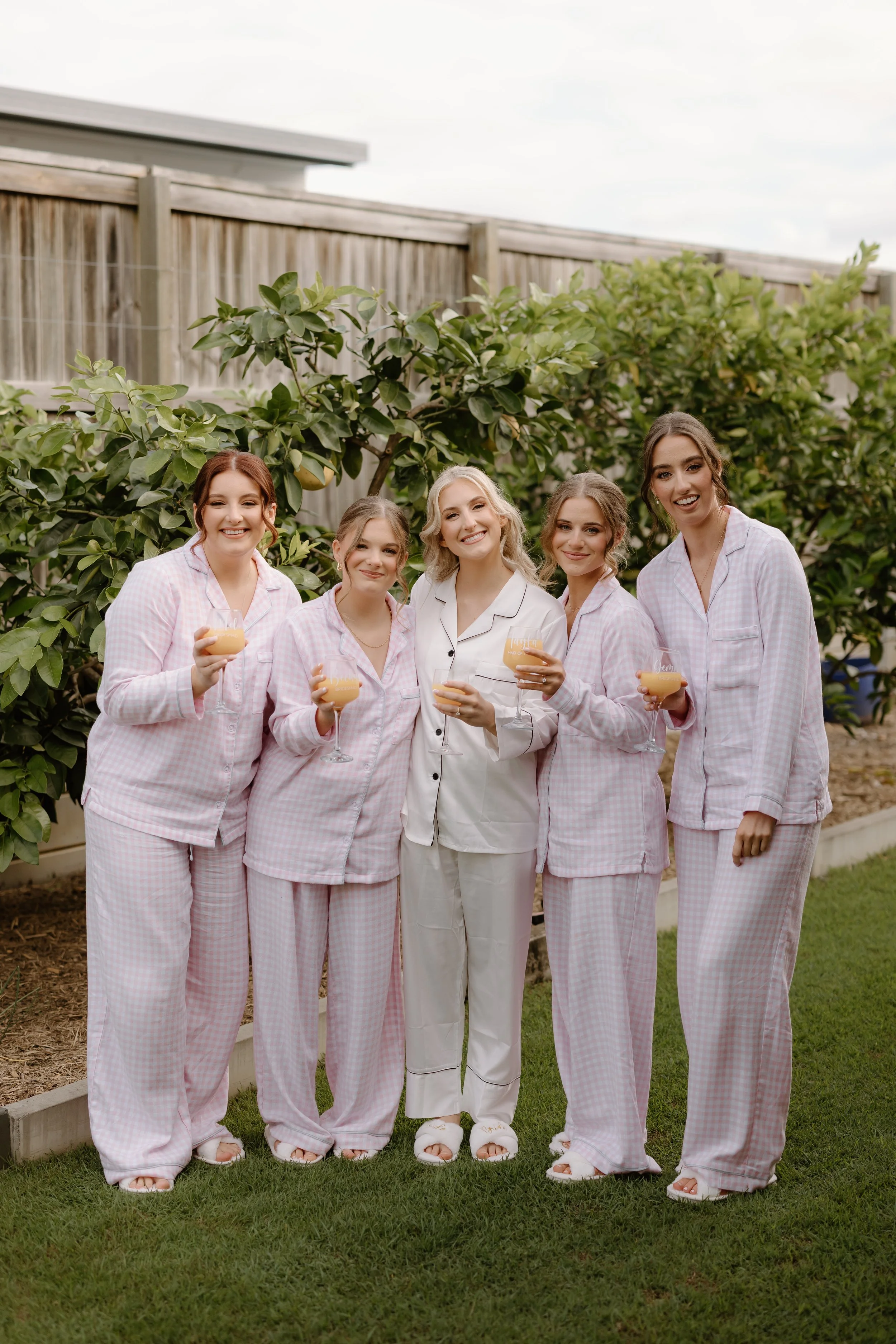 Five women in pajamas standing outdoors holding drinks, smiling, with a garden and wooden fence in the background.