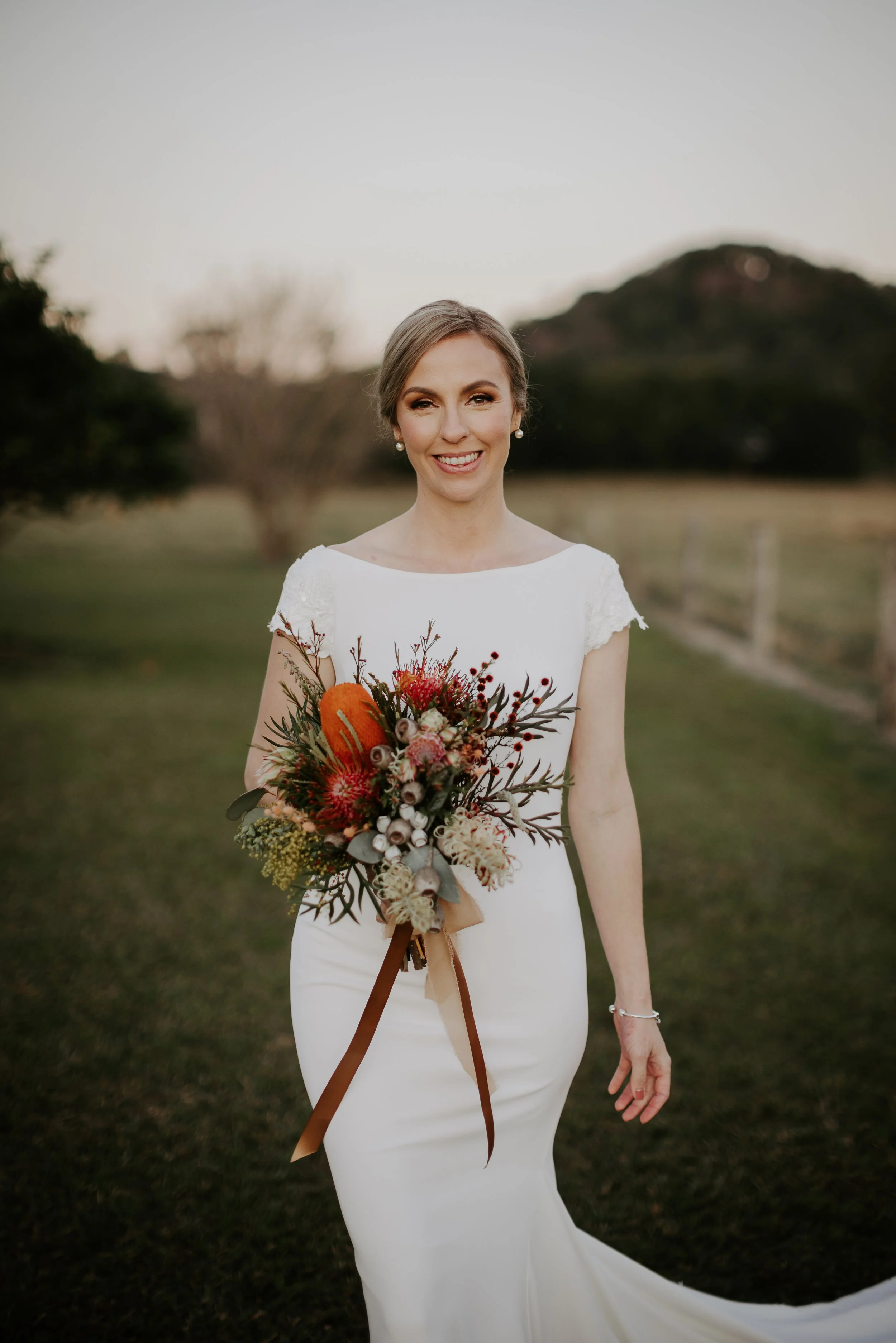 Bride in white wedding dress holding a bouquet outdoors at sunset, smiling.