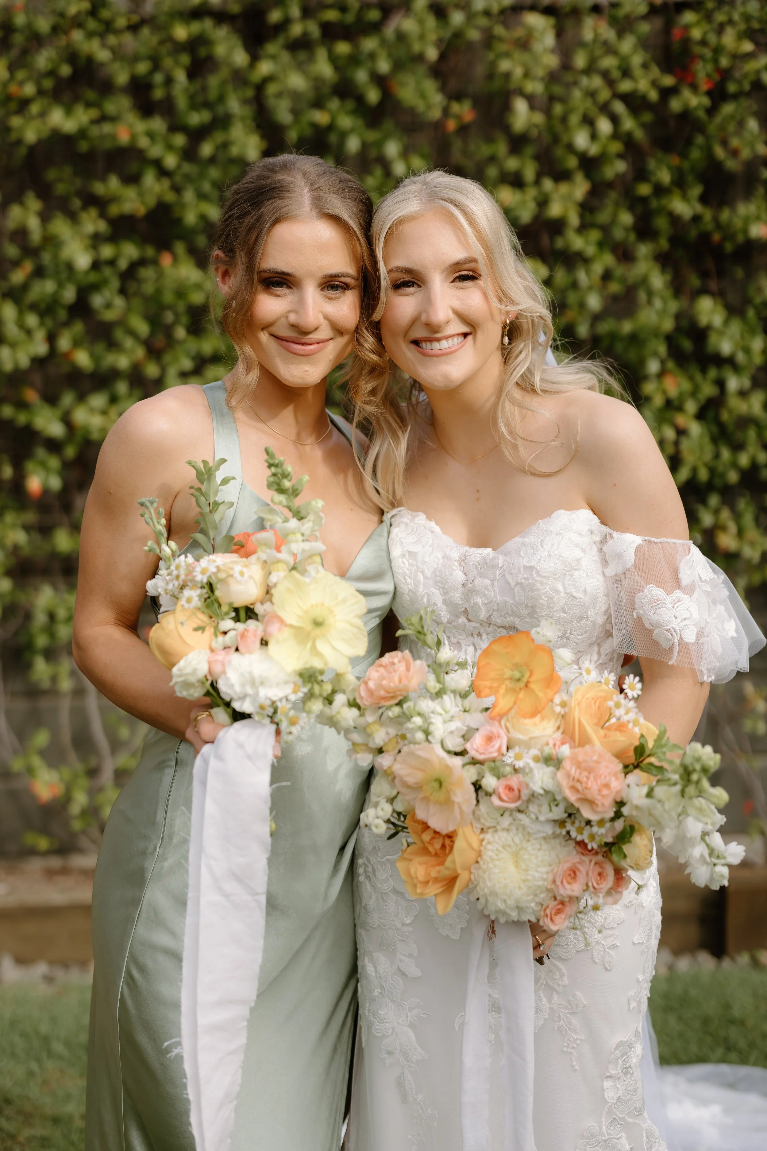 Two women in wedding attire holding bouquets, smiling outdoors with green foliage in the background.