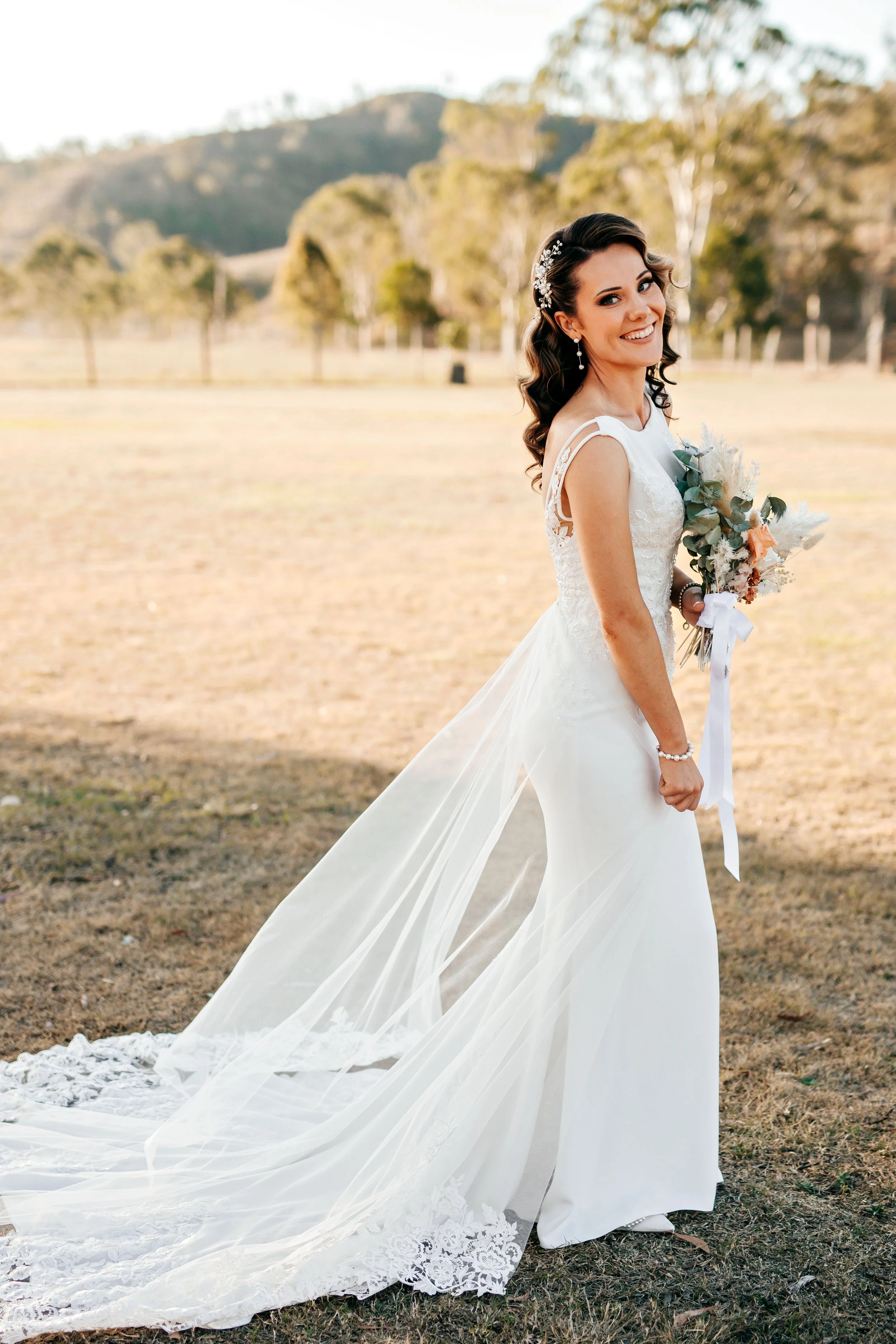 A bride in a white wedding dress standing outdoors in a field, holding a bouquet, smiling at the camera with mountains and trees in the background.