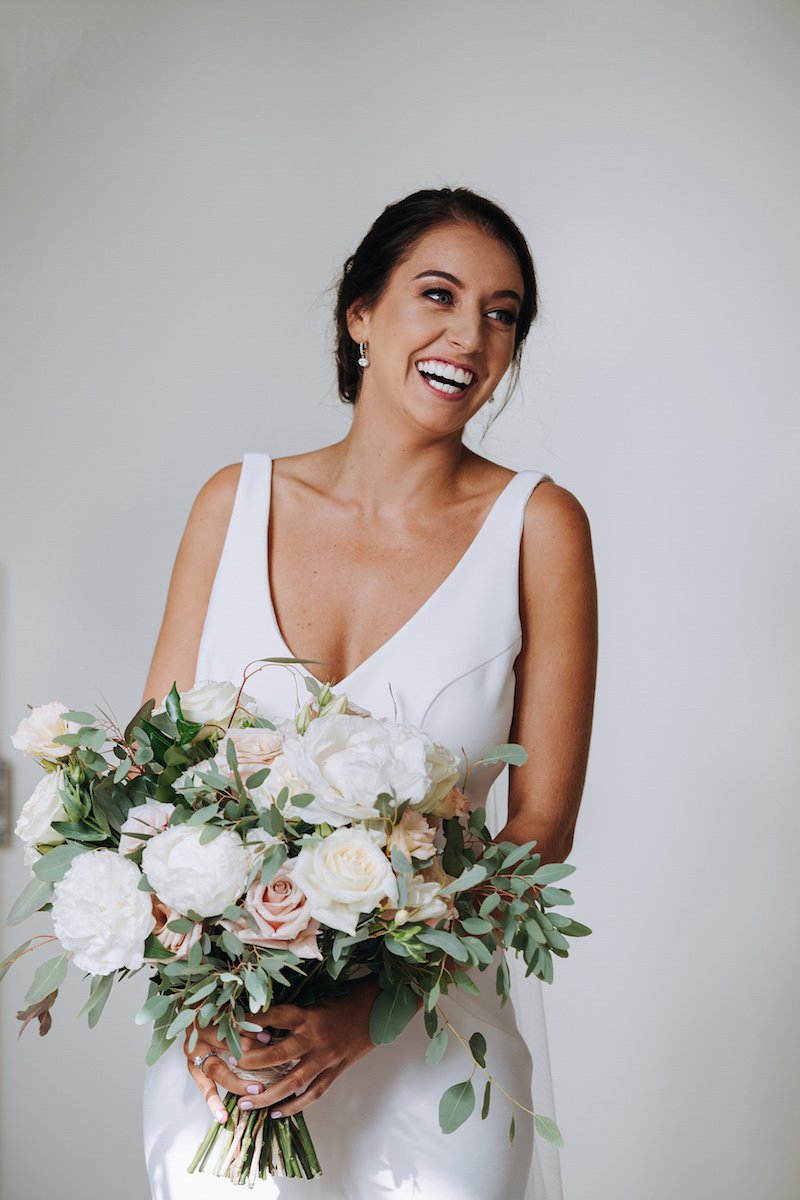 A smiling woman in a white dress holding a bouquet of white and pale pink flowers.