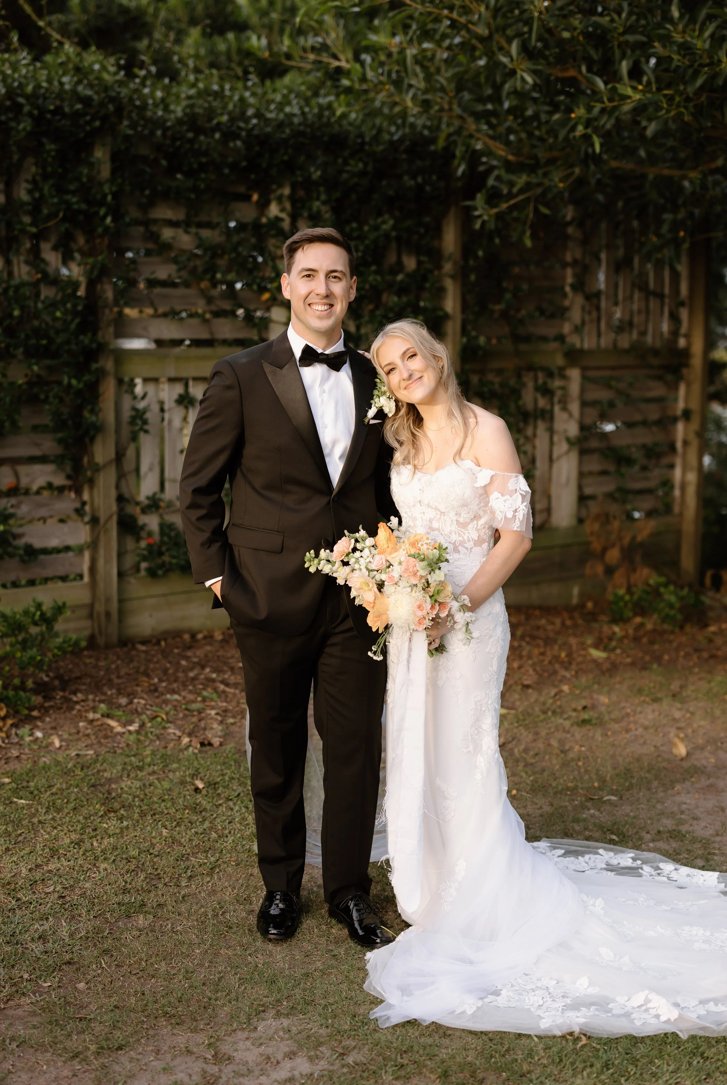 A newlywed couple standing outdoors, the groom in a black tuxedo with a bow tie, and the bride in a white lace wedding dress holding a bouquet of flowers, smiling.