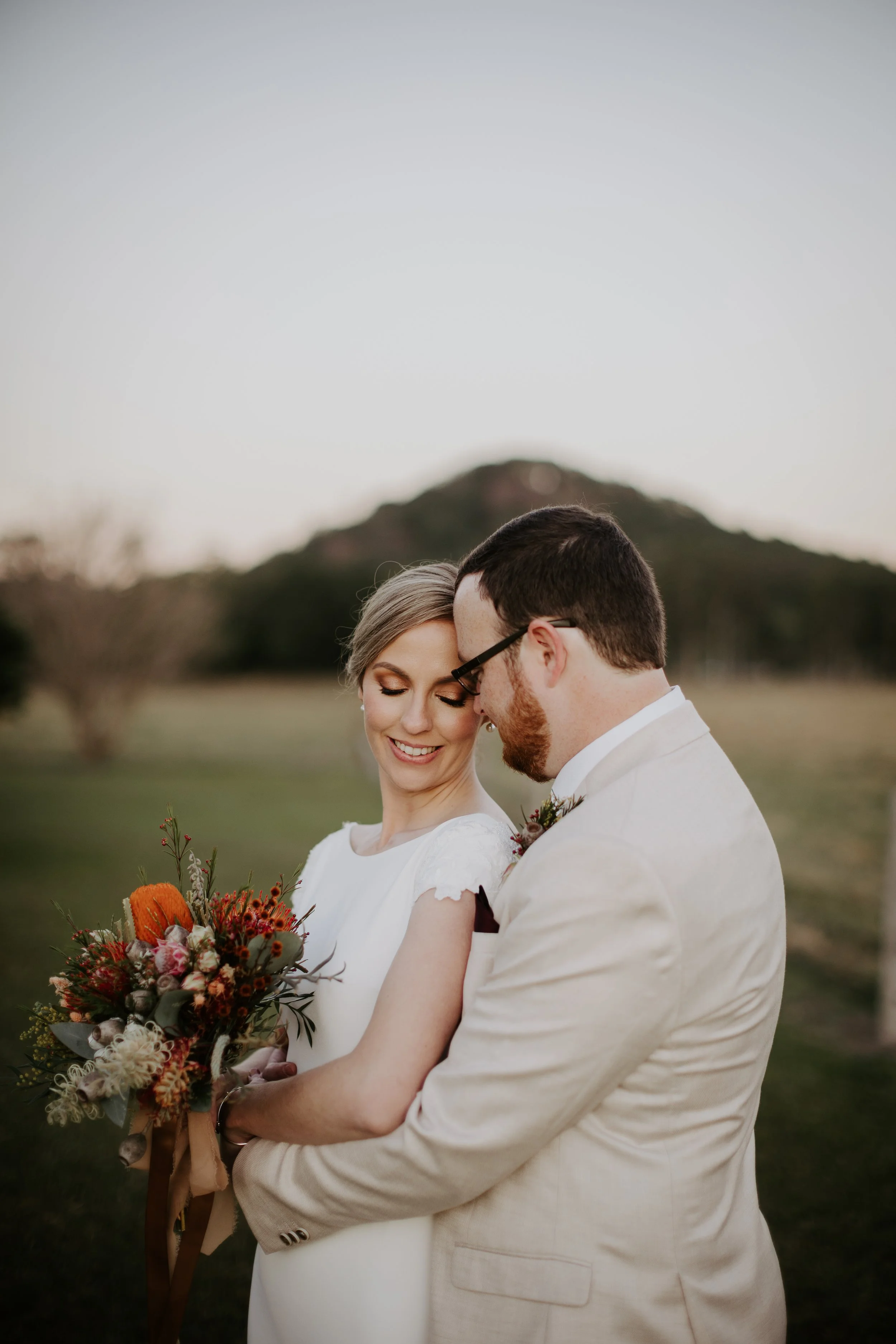 A bride and groom embrace outdoors during sunset, with a mountain in the background. The bride holds a bouquet of flowers, and both are smiling with eyes closed.