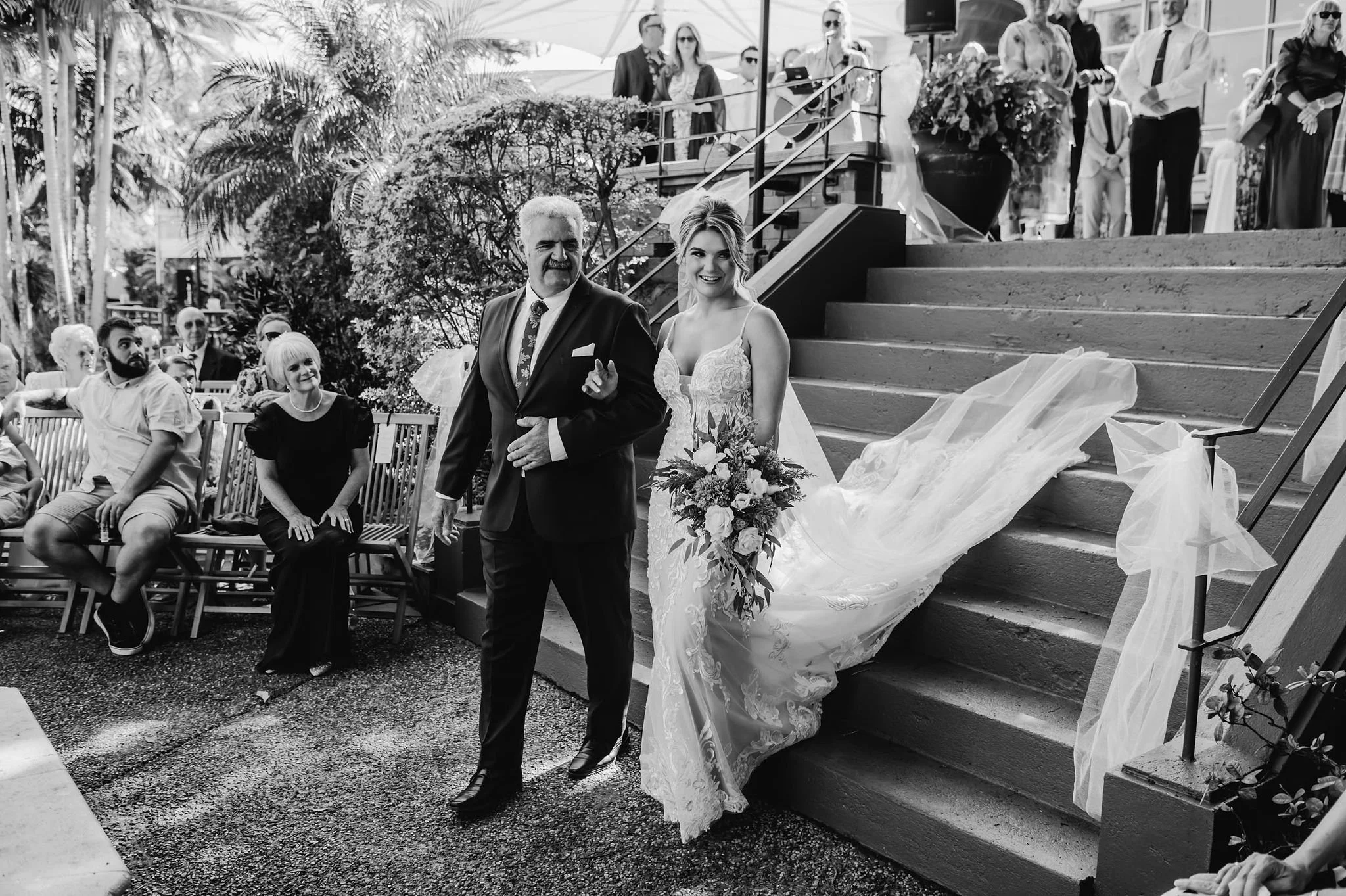 A bride in a wedding gown holding a bouquet, walking down the stairs accompanied by a man in a suit at a wedding ceremony