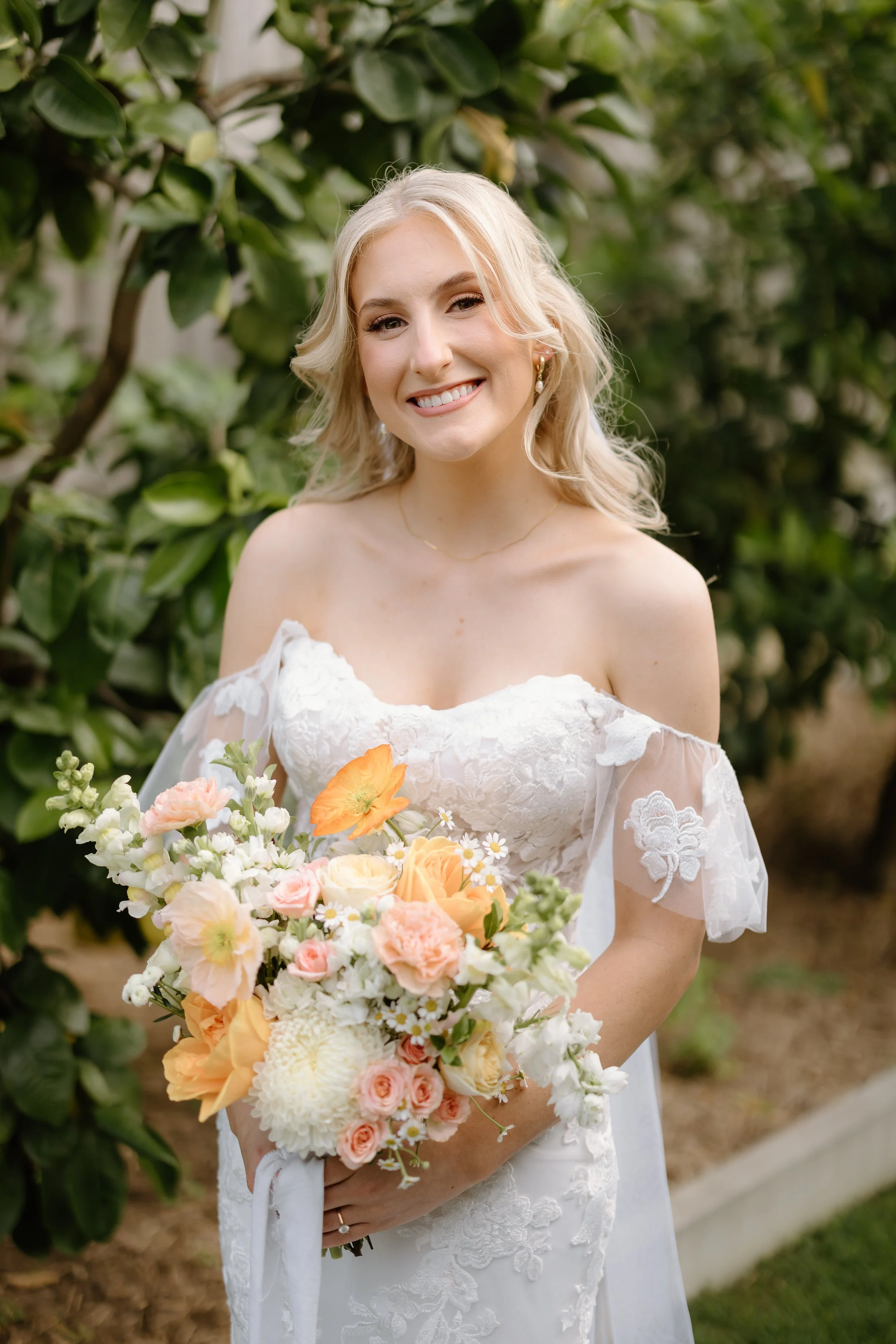 A bride with blonde hair in loose waves, wearing a white off-shoulder lace wedding dress, holding a bouquet of peach, white, and pink flowers, smiling outdoors with greenery in the background.