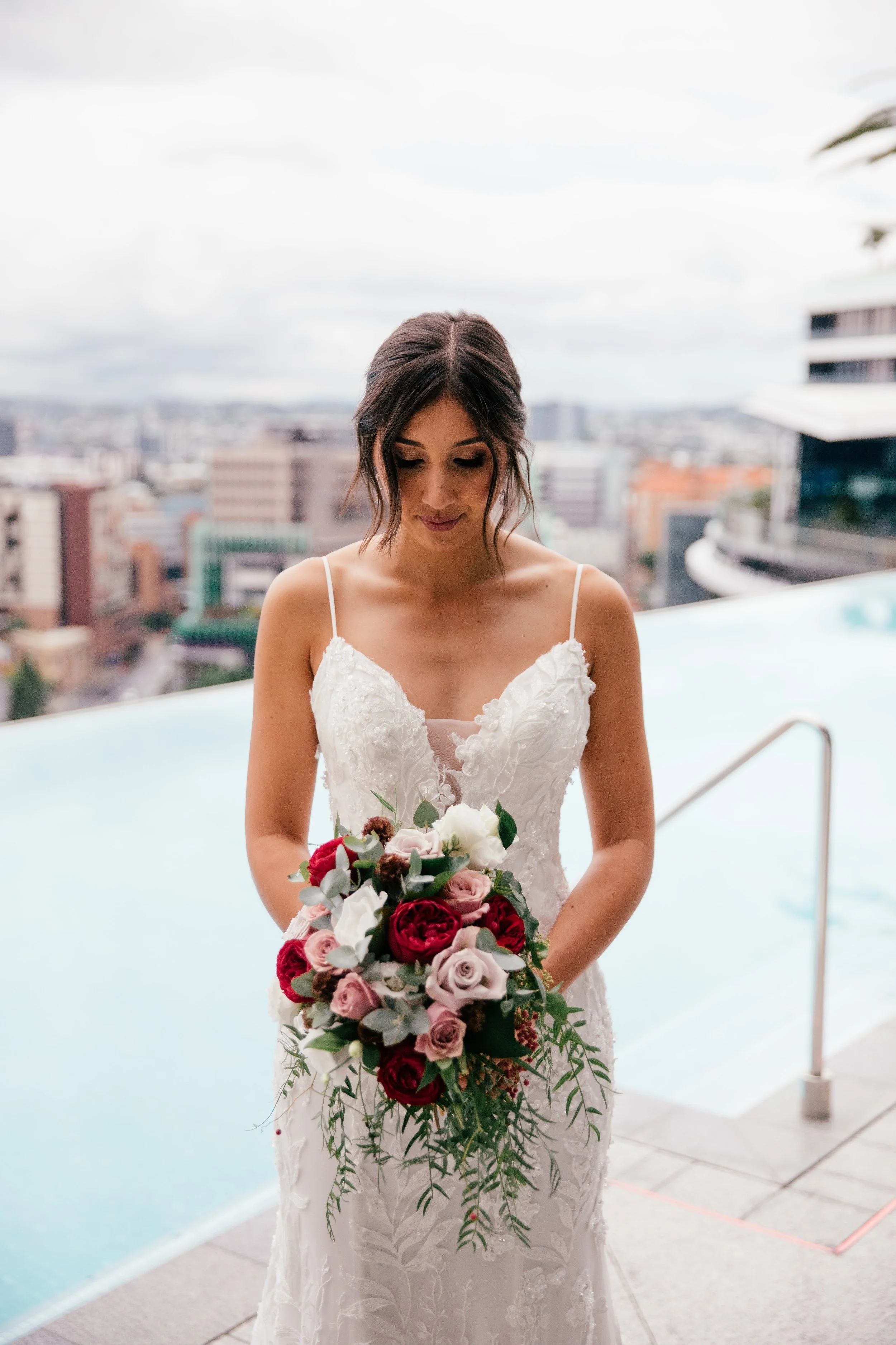 A woman in a white bridal gown holding a bouquet of red, pink, and white roses with greenery, standing near a rooftop pool with a city skyline in the background.
