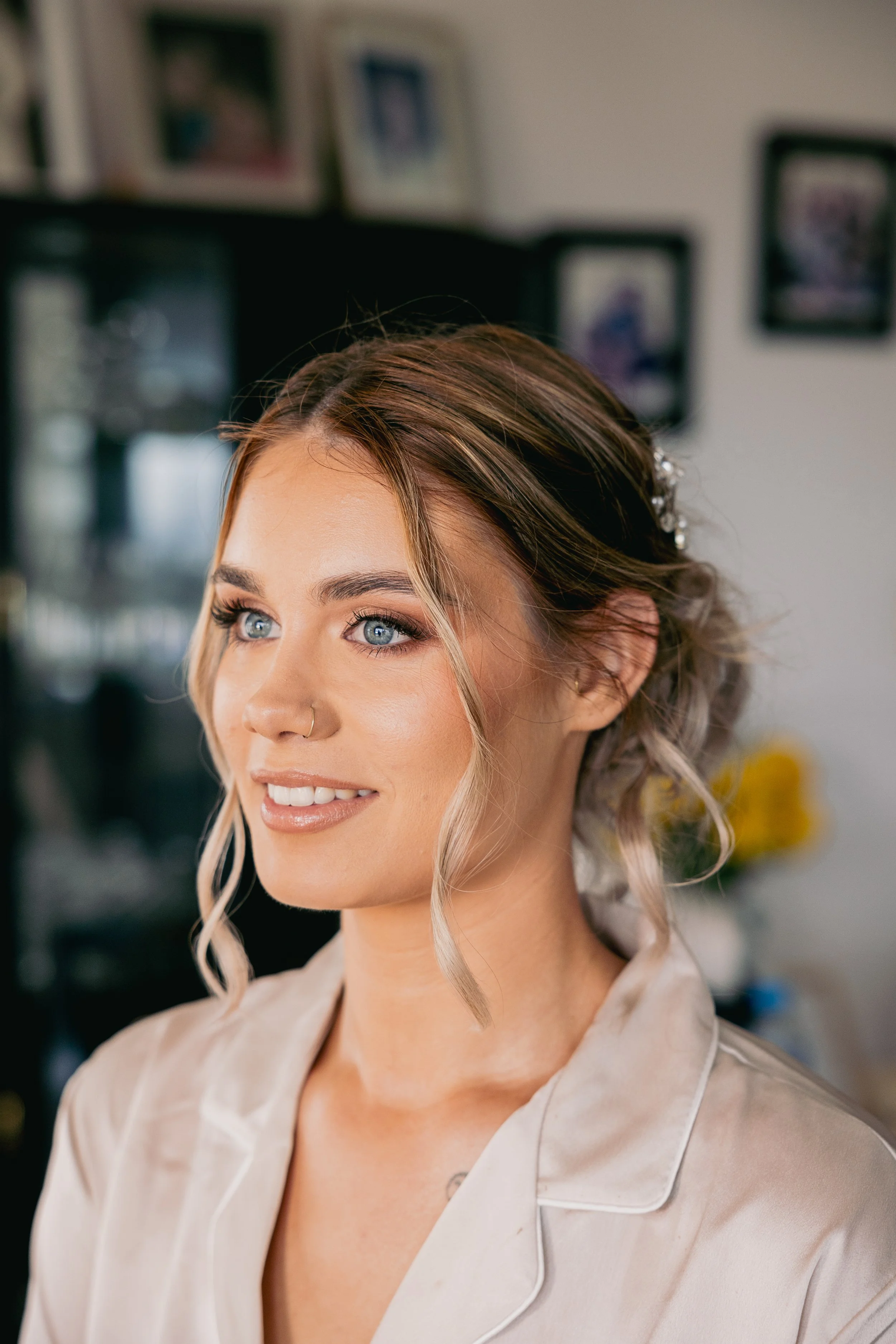 A smiling woman with blonde hair styled in loose curls, wearing light makeup, and a satin beige robe. She has a nose ring and a hair accessory, and is in a room with framed pictures and a blurred background.