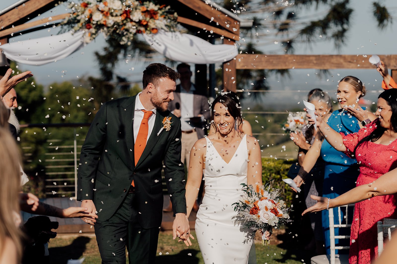 Bride and groom walking hand in hand among guests throwing confetti during outdoor wedding ceremony.
