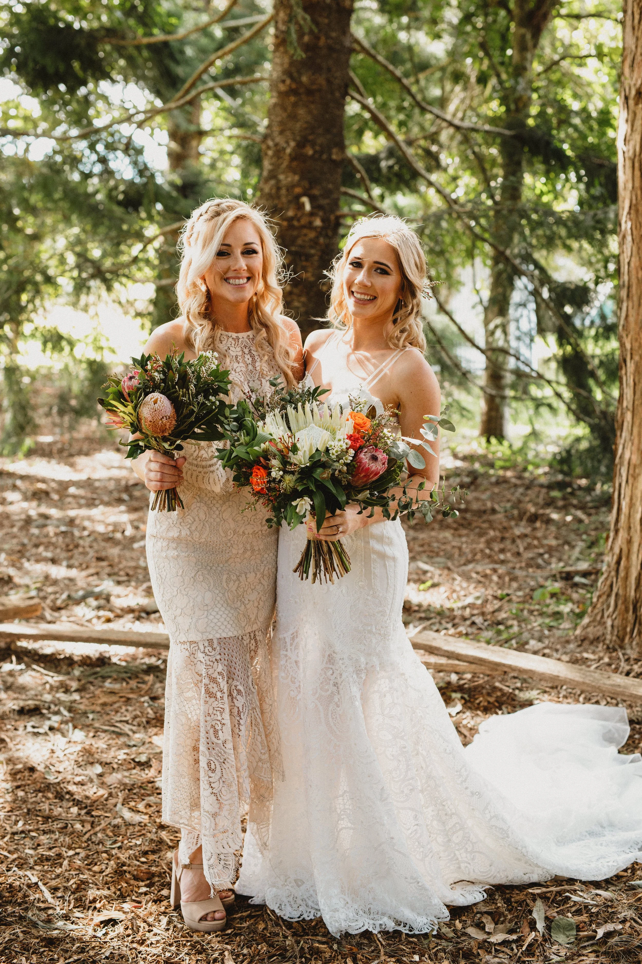 Two women in wedding dresses holding bouquets, standing outdoors in a wooded area.