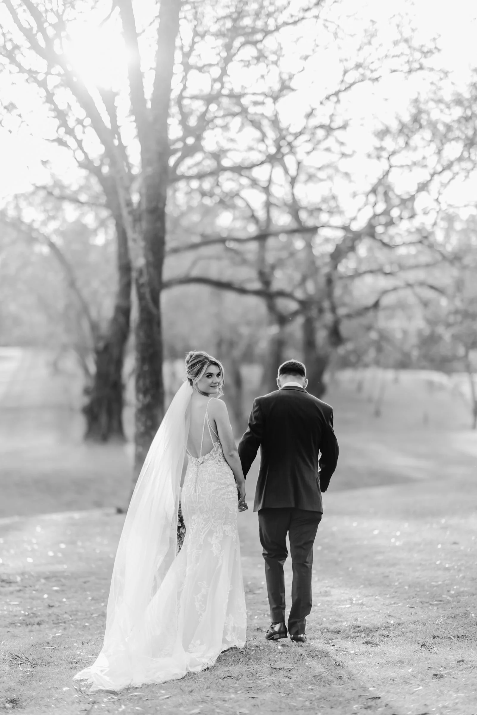 Black-and-white photo of a bride and groom walking hand in hand in a park, with trees and sunlight in the background.