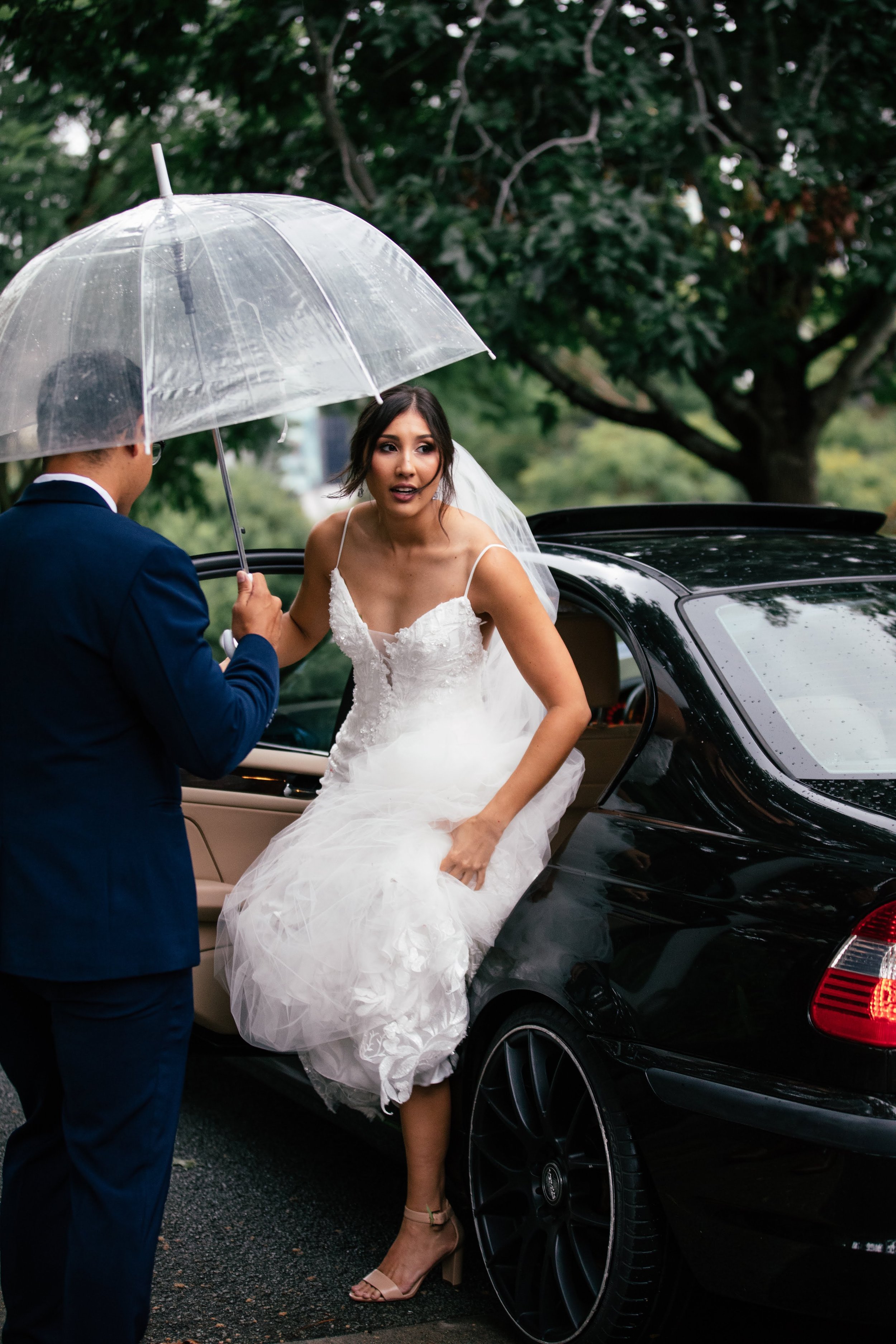Bride in wedding dress getting into a black luxury car while holding an umbrella, with a man in a suit assisting her, in a rainy outdoor setting with trees in the background.