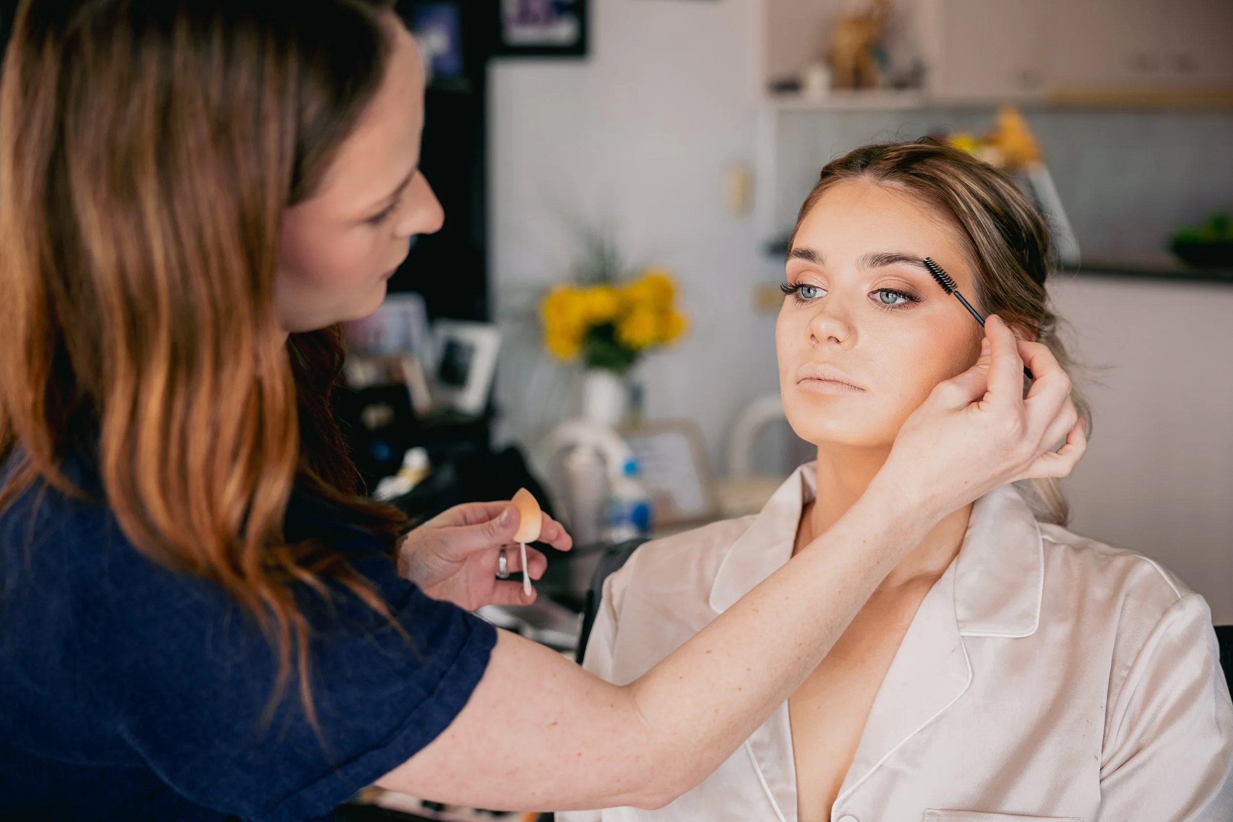 Makeup artist applying makeup to a woman with light skin and blue eyes, using a spoolie brush to groom her eyebrows. The woman is wearing a satin robe and is looking slightly to the side. The background shows a kitchen with flowers on the counter.