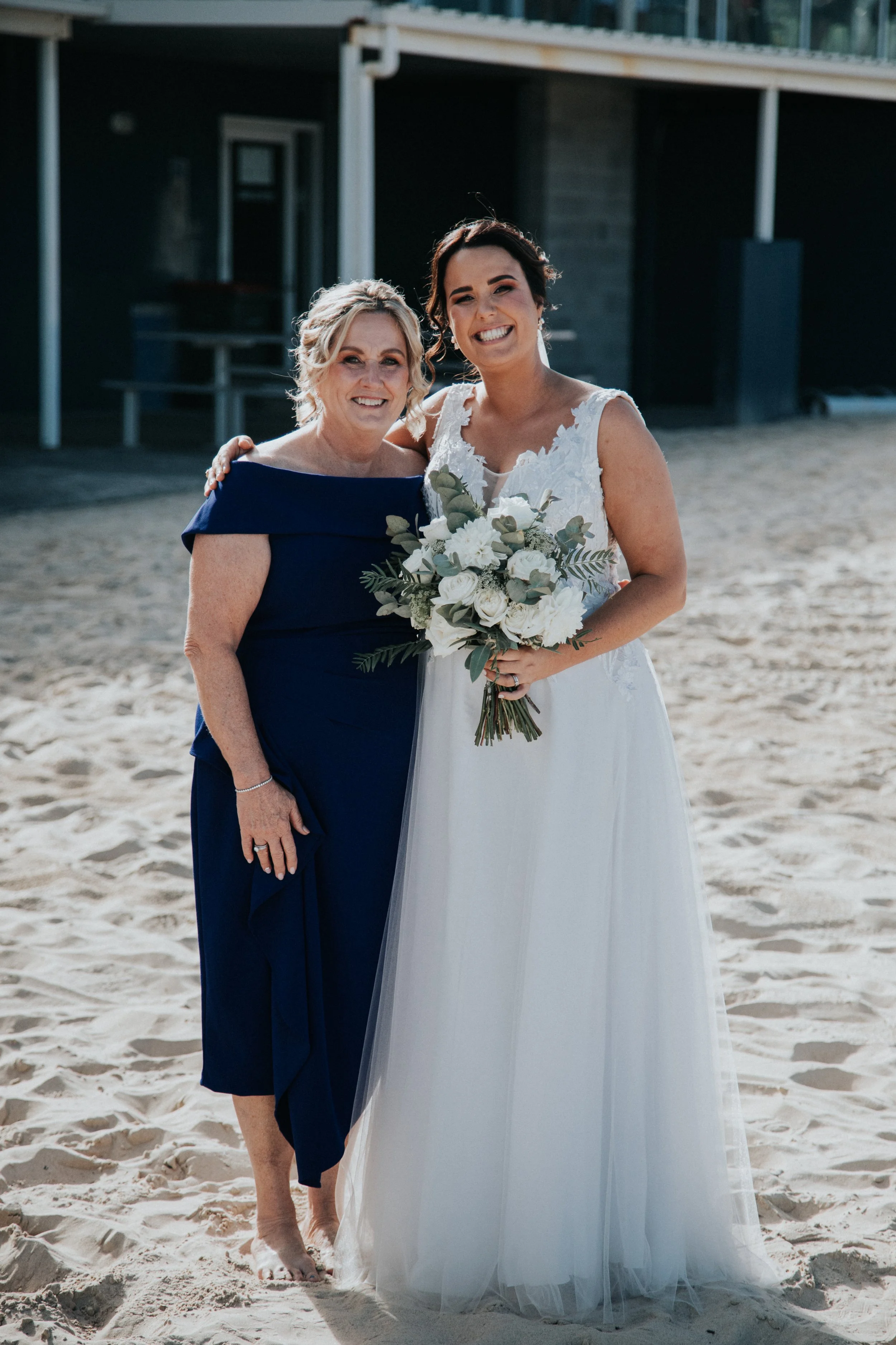 A bride in a white dress holding a bouquet, standing on a sandy beach with an older woman in a dark blue dress, both smiling and embracing.