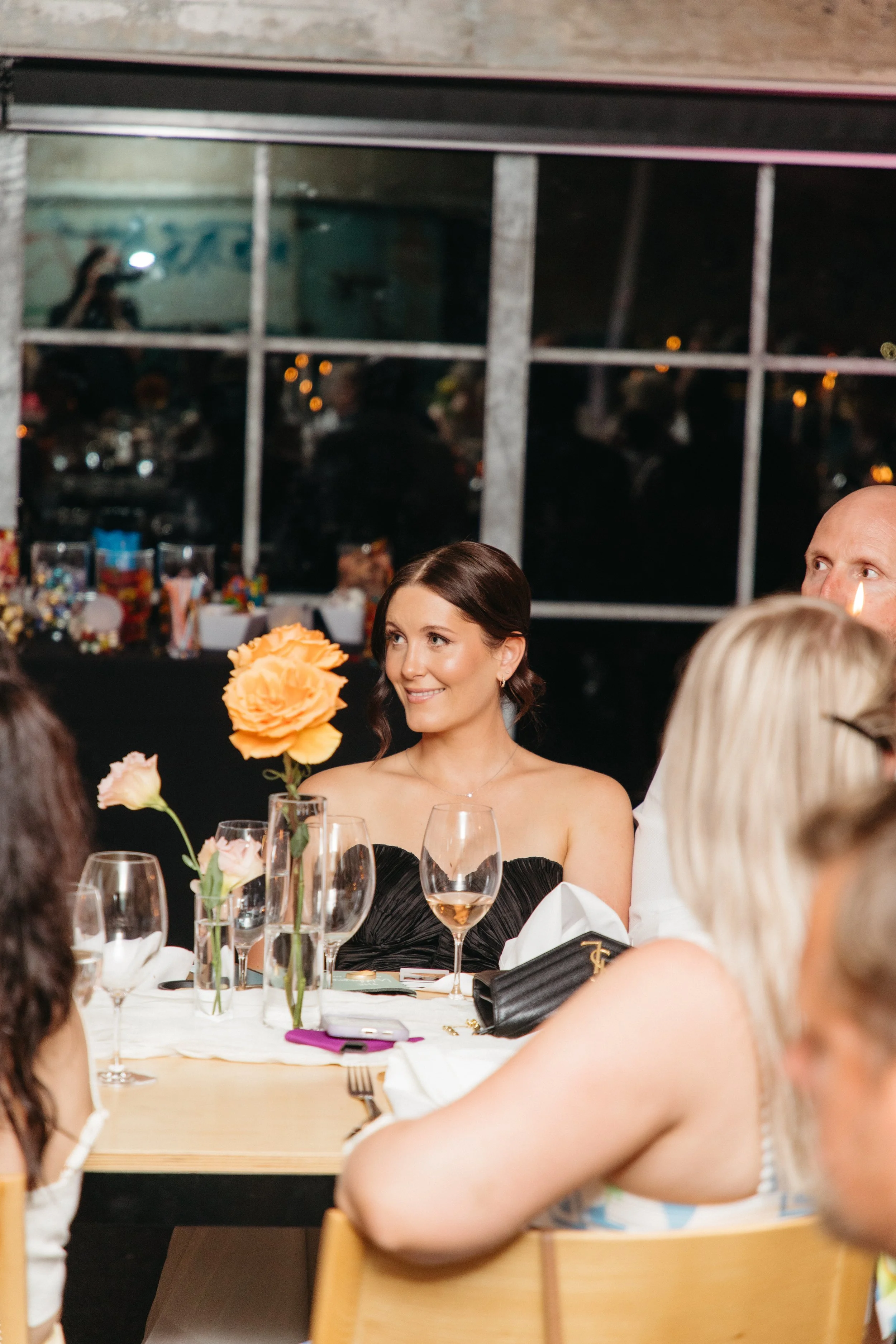 A woman with dark brown hair in an updo, wearing a black strapless dress, sitting at a dinner table with floral centerpieces, glasses, and table settings during a social event or wedding reception at night.