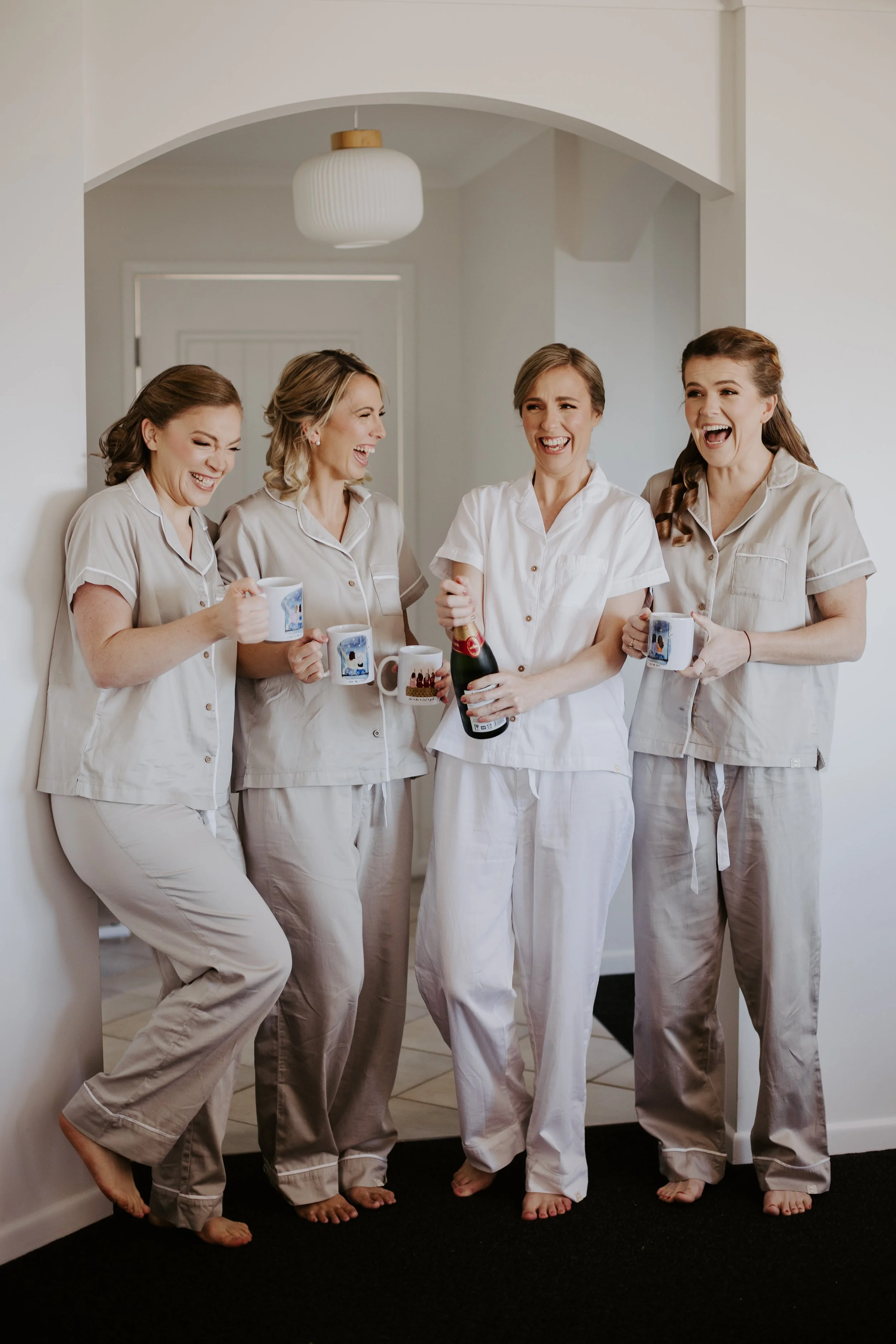 Group of five women in pajamas laughing and holding cups and a bottle in a cozy home setting.