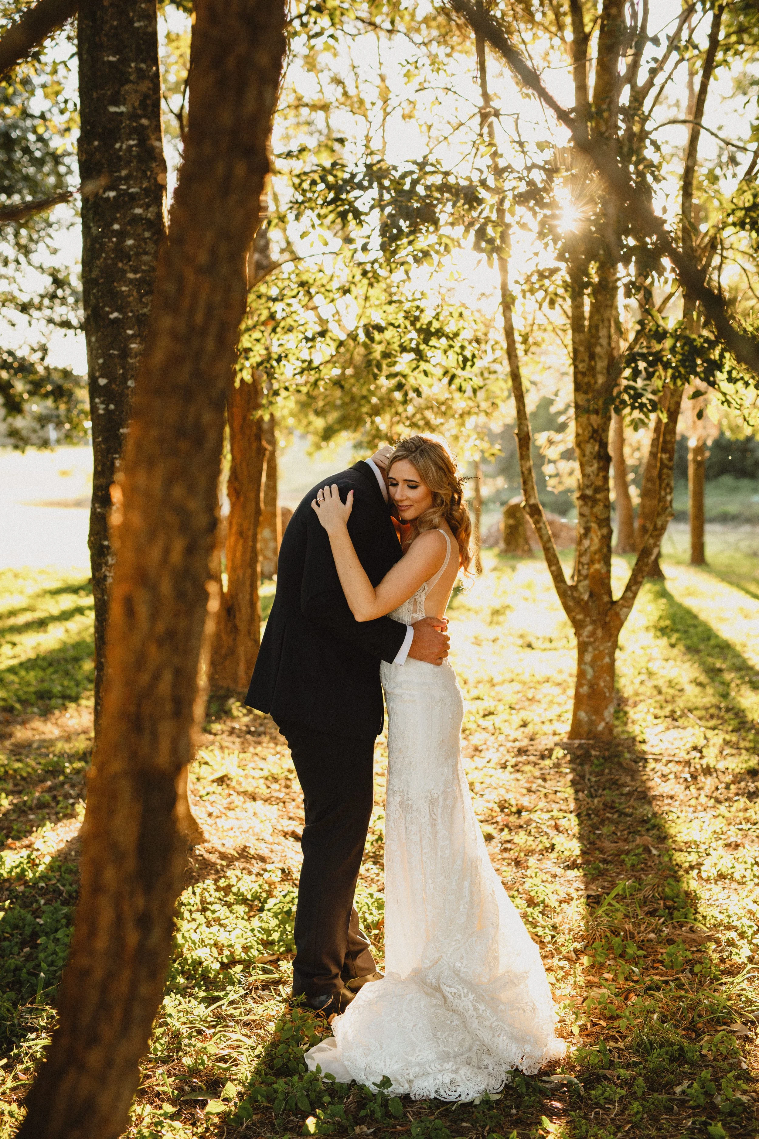 A bride and groom share a tender embrace in a wooded outdoor setting during sunset.
