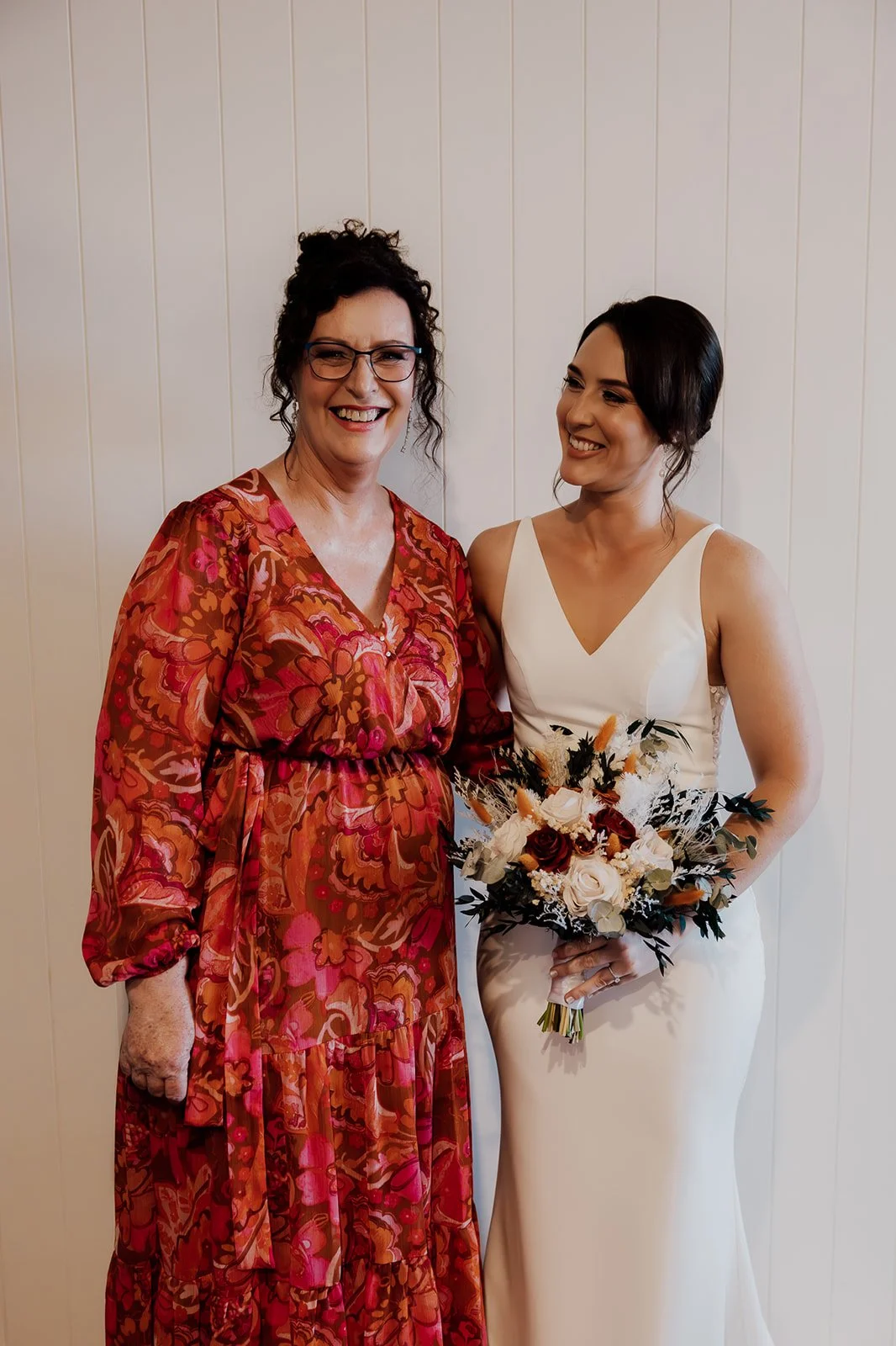 A woman in a floral dress standing next to a bride in a white gown holding a bouquet of flowers, both smiling for a photo in front of a wooden wall.
