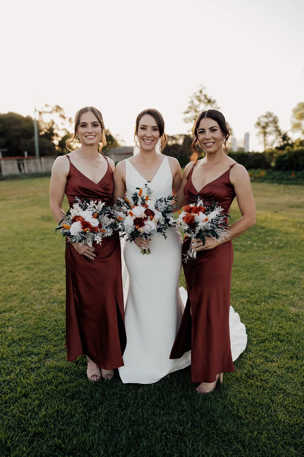 Three women in formal dresses standing outdoors on grass, holding bouquets of flowers, smiling at the camera during sunset.