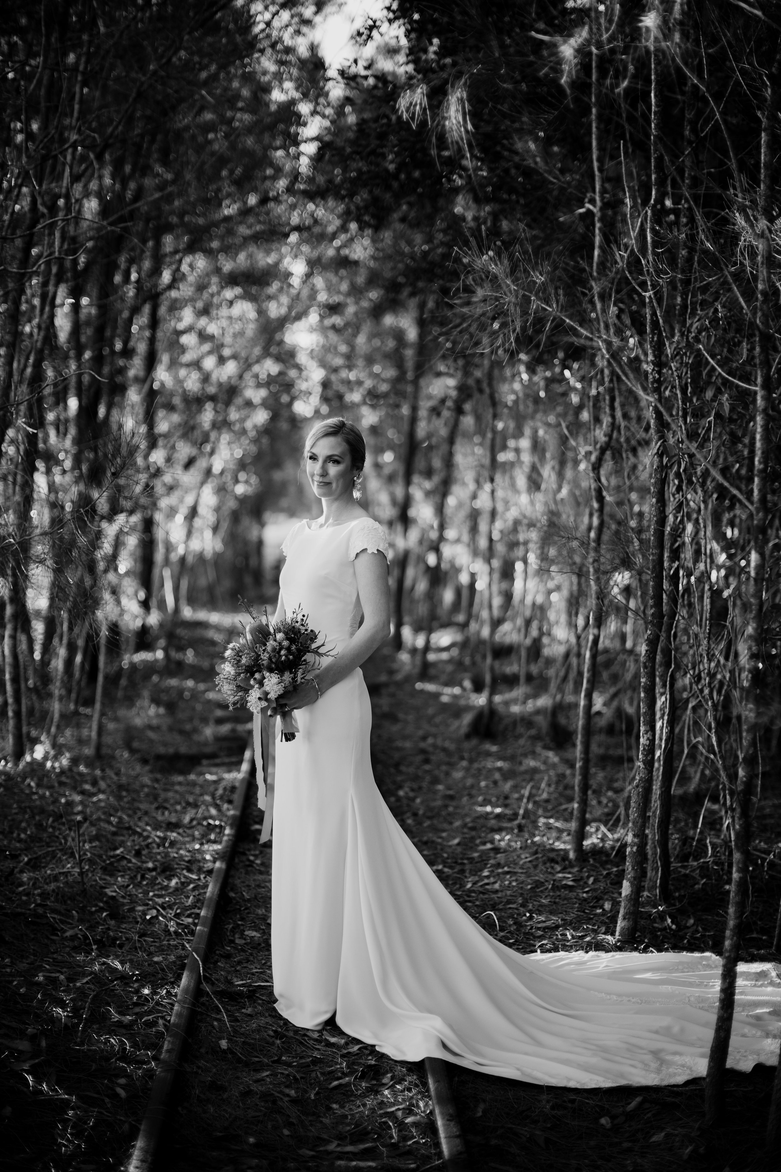 Black and white photo of a bride in a wedding dress holding a bouquet standing on a forest path.
