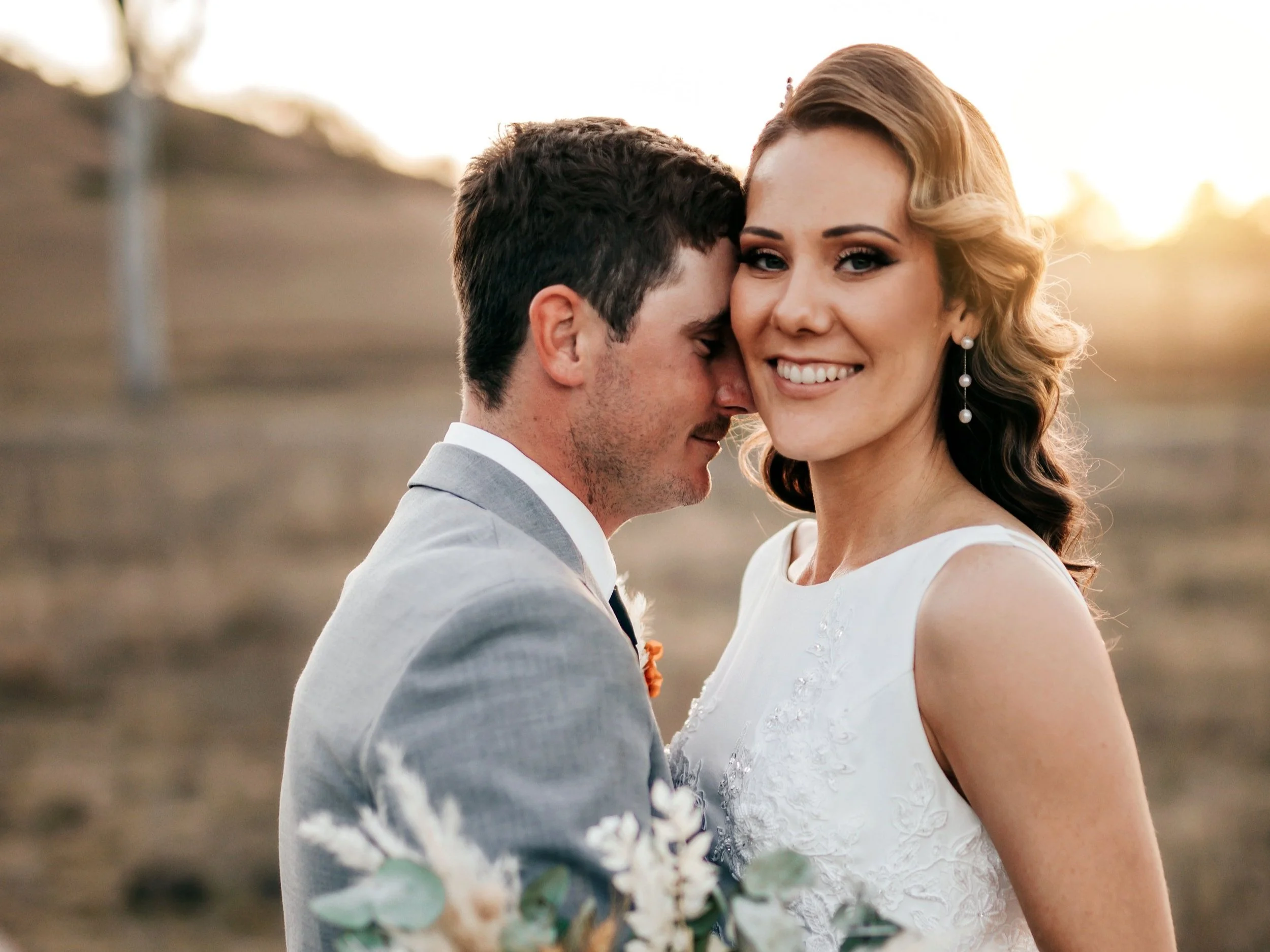 A bride and groom sharing a close, intimate moment outdoors at sunset, with the groom softly pressing his forehead against the bride's temple, smiling warmly.