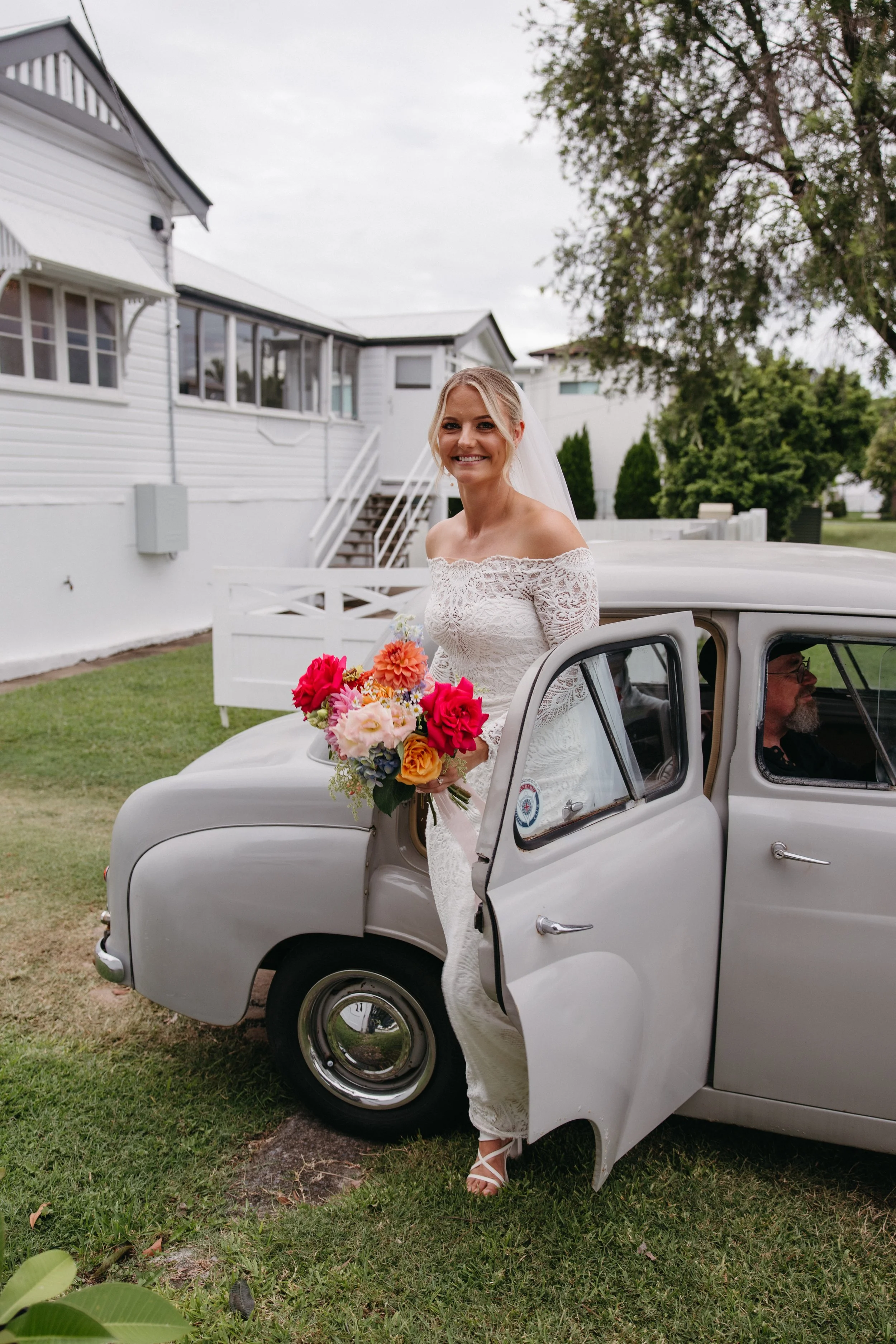 A bride in a white lace wedding dress holding a colorful bouquet of flowers, smiling as she stands outside a vintage silver car with the door open, on a green lawn with white houses and trees in the background.