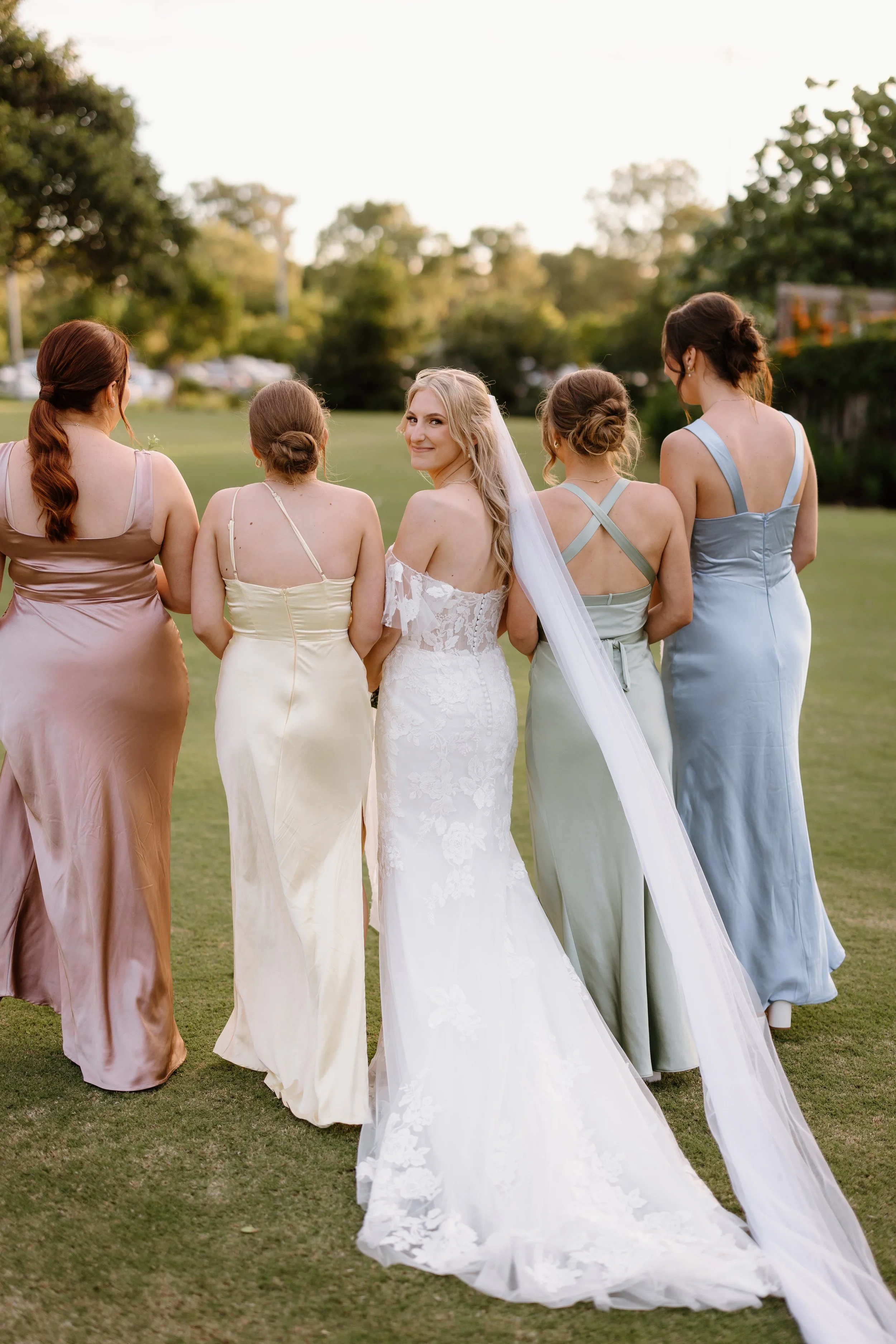 Bride in white lace wedding dress with long veil and four bridesmaids in pastel-colored dresses walking outdoors on grass field during sunset.