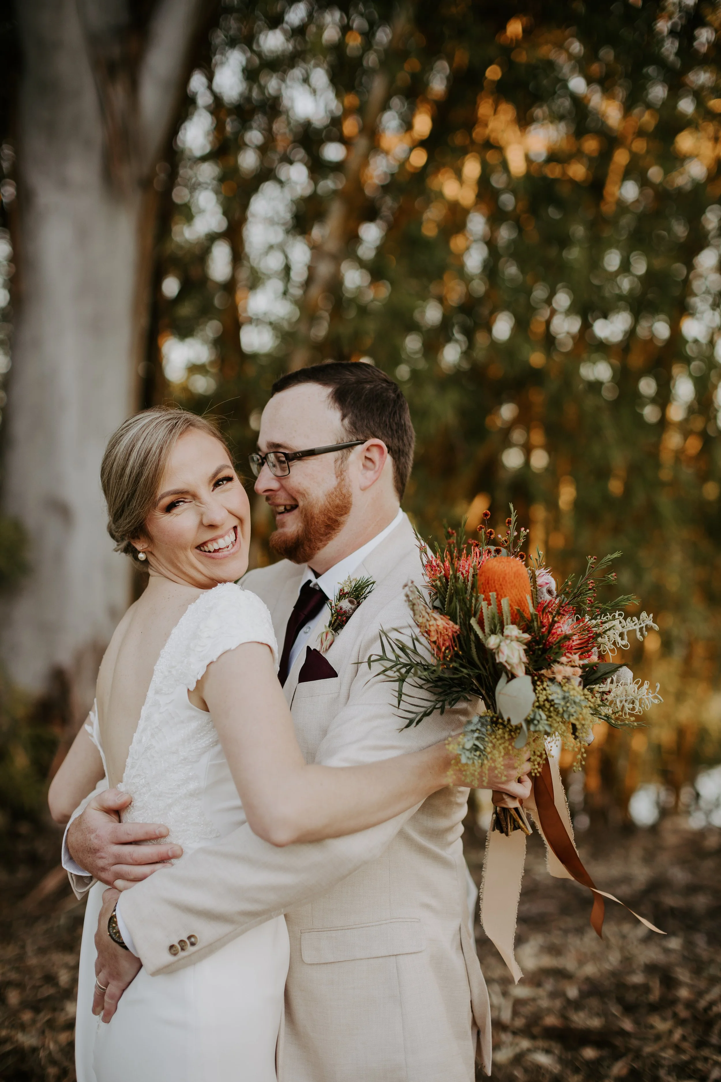 A happy bride and groom embracing outdoors, smiling at each other, with the bride holding a colorful bouquet. The background features trees with autumn leaves.