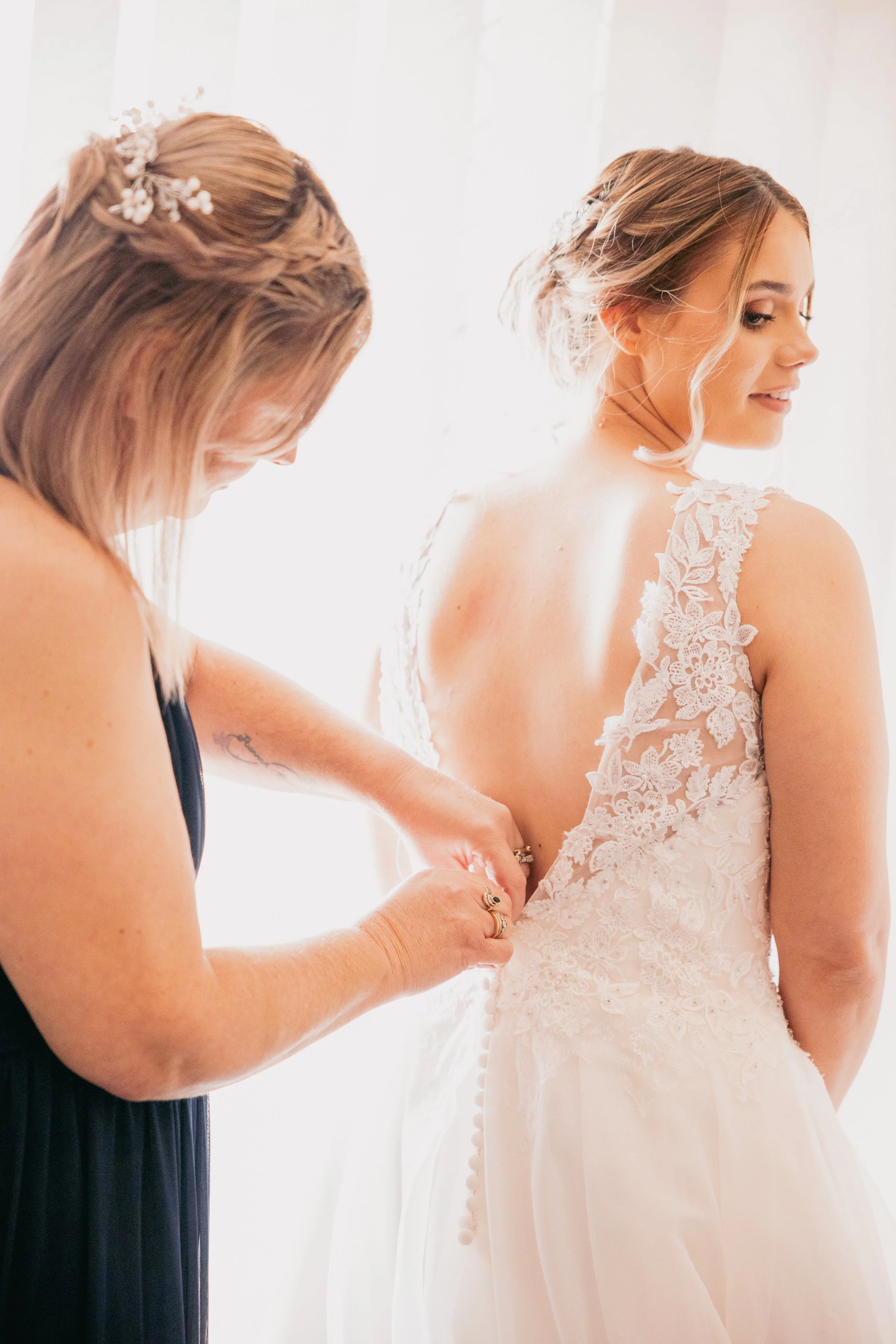 A bride getting help with her wedding dress from a woman. The bride is wearing a lace wedding gown, and the woman is adjusting the back of the dress.