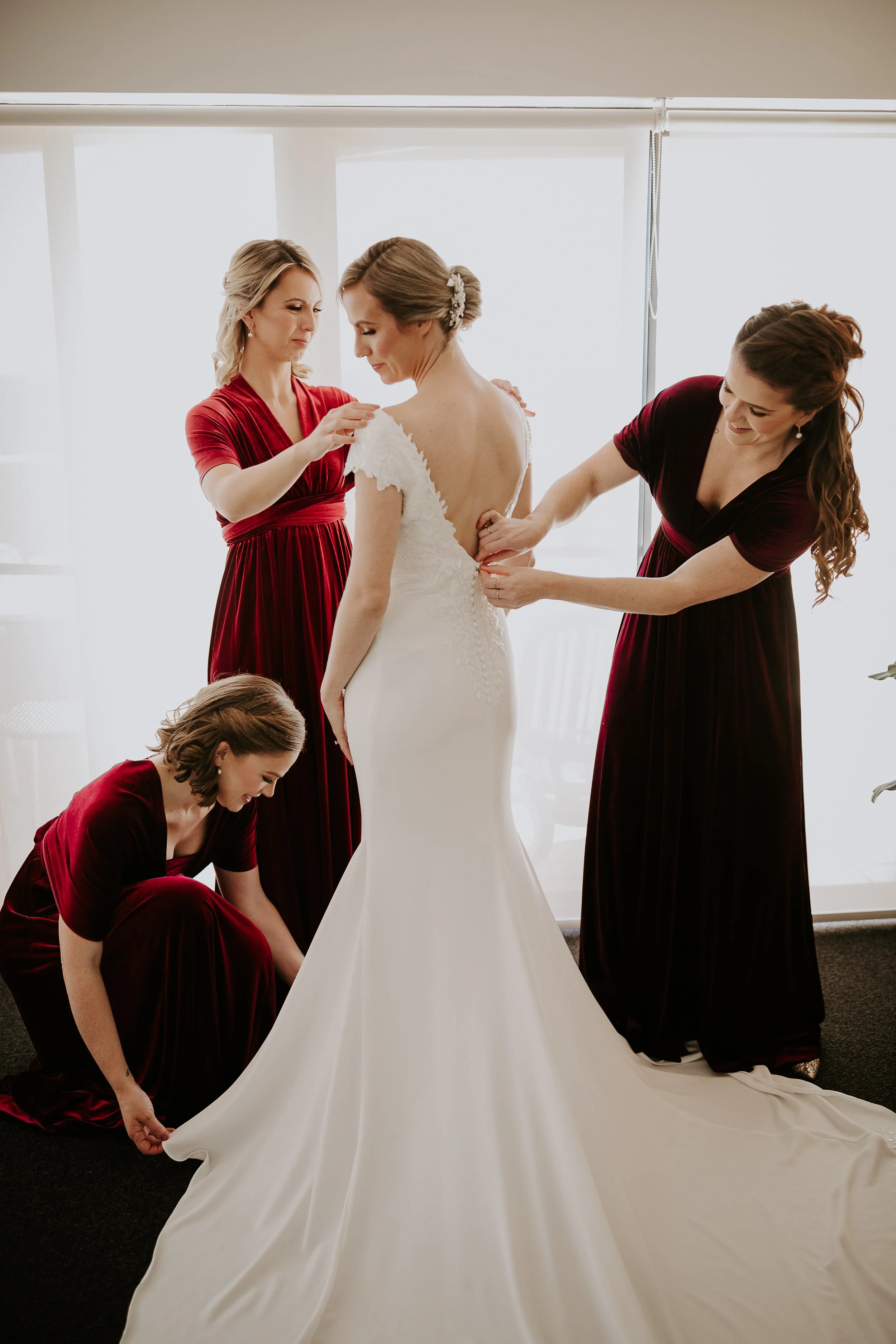 Four women, dressed in red and black gowns, helping a bride in a white wedding dress with back buttons, as they prepare for her wedding in front of a brightly lit window.