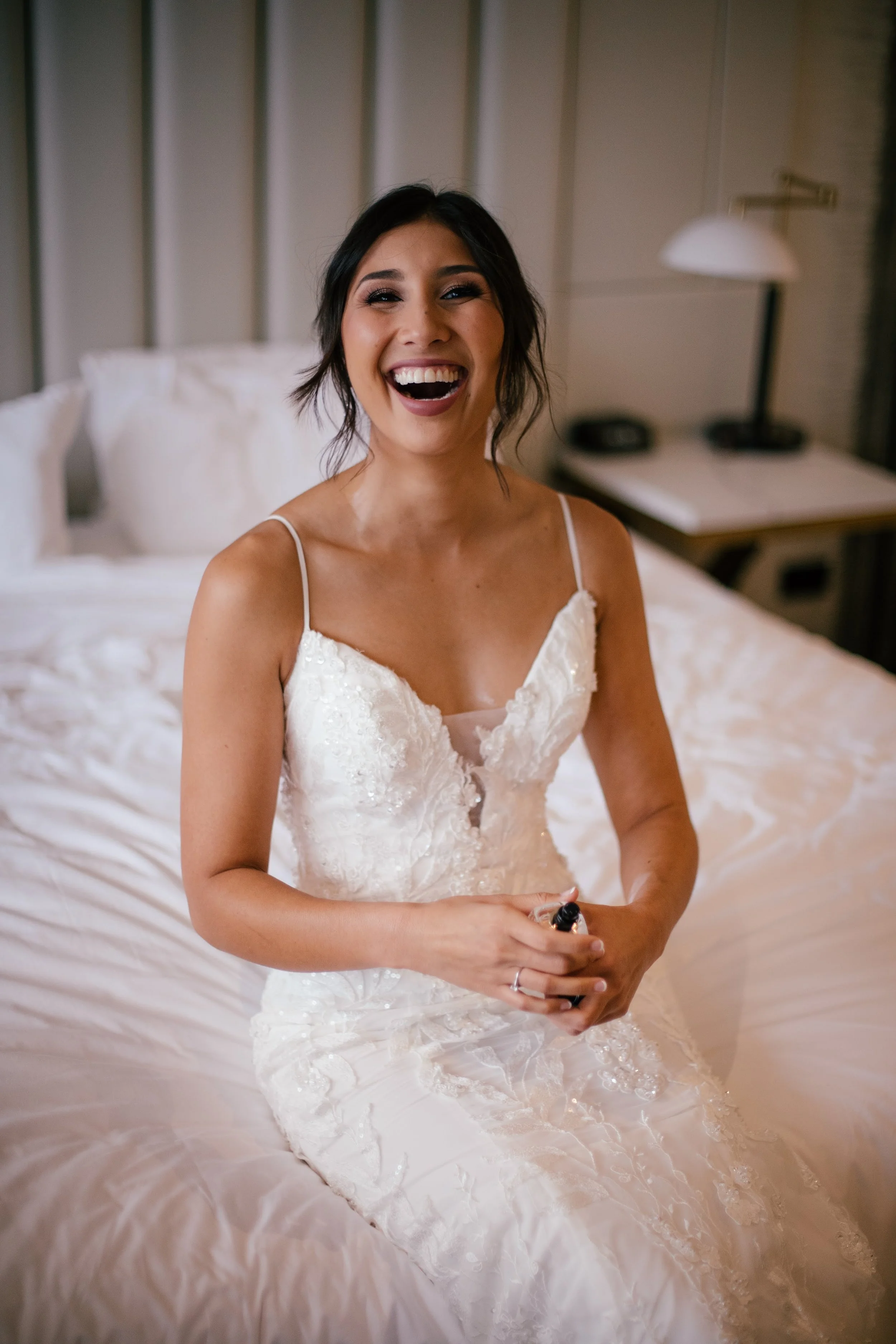 A woman in a white wedding dress sitting on a bed, smiling and laughing in a hotel room.