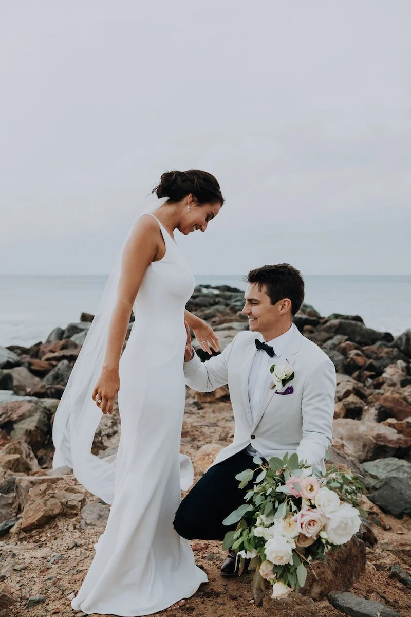 A bride and groom at the beach, with the groom kneeling on one knee holding a bouquet of flowers, and the bride smiling down at him against a rocky shoreline and overcast sky.