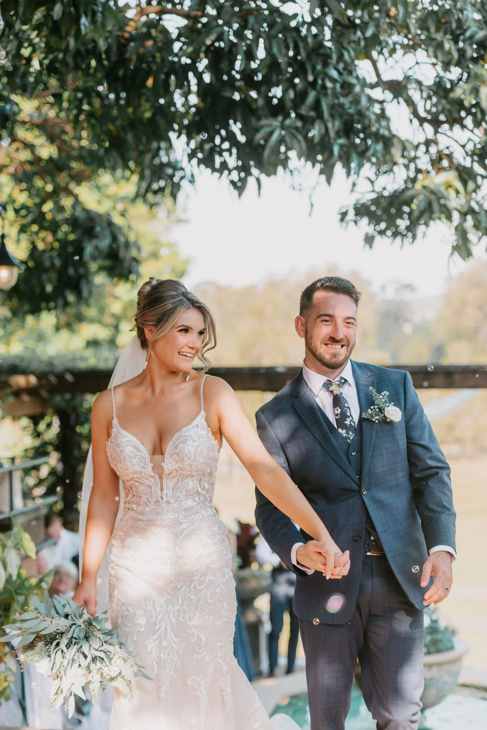 A bride and groom happily walking hand in hand outdoors during their wedding celebration, smiling and dressed in formal wedding attire.