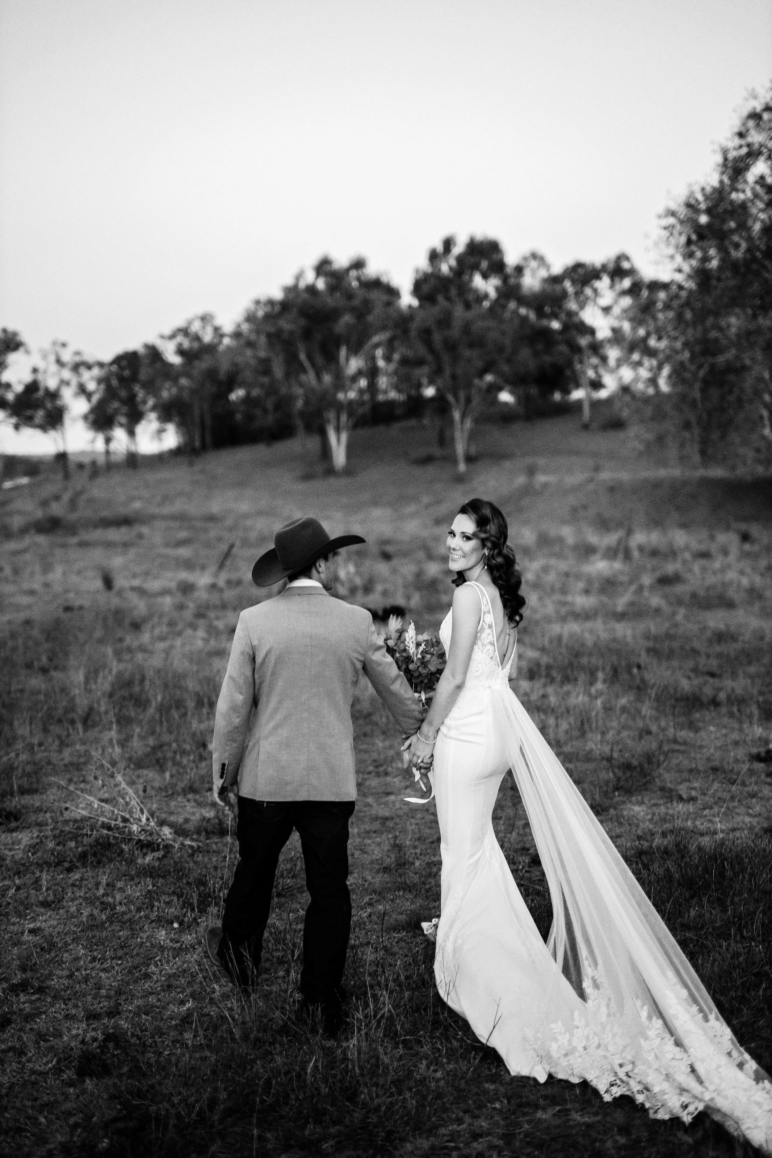 Black and white photo of a bride in a wedding dress holding hands with a man in a suit and cowboy hat, standing in an open field with trees in the background.