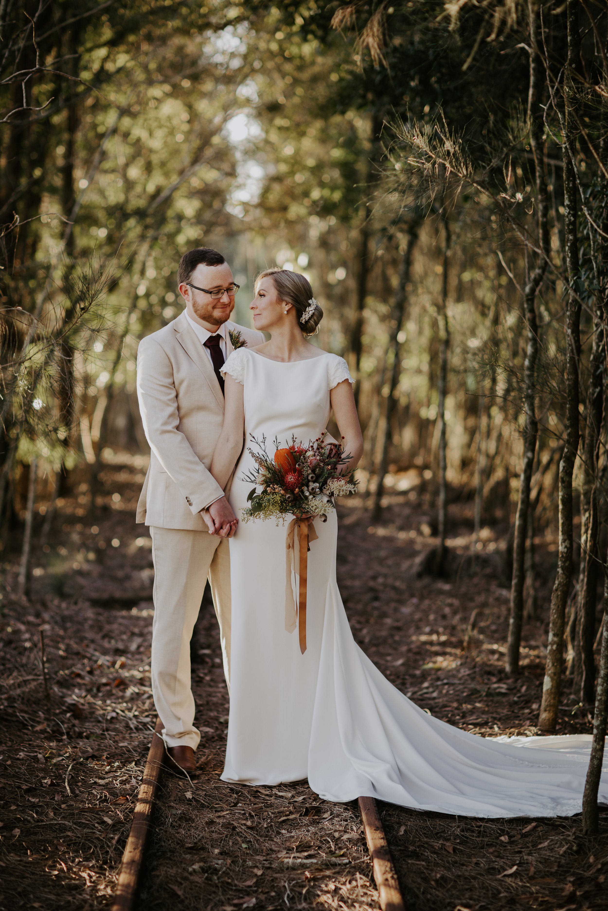 A bride and groom standing on train tracks in a wooded area during their wedding, holding hands and gazing at each other. The bride is wearing a white wedding dress and holding a bouquet, and the groom is in a light beige suit.