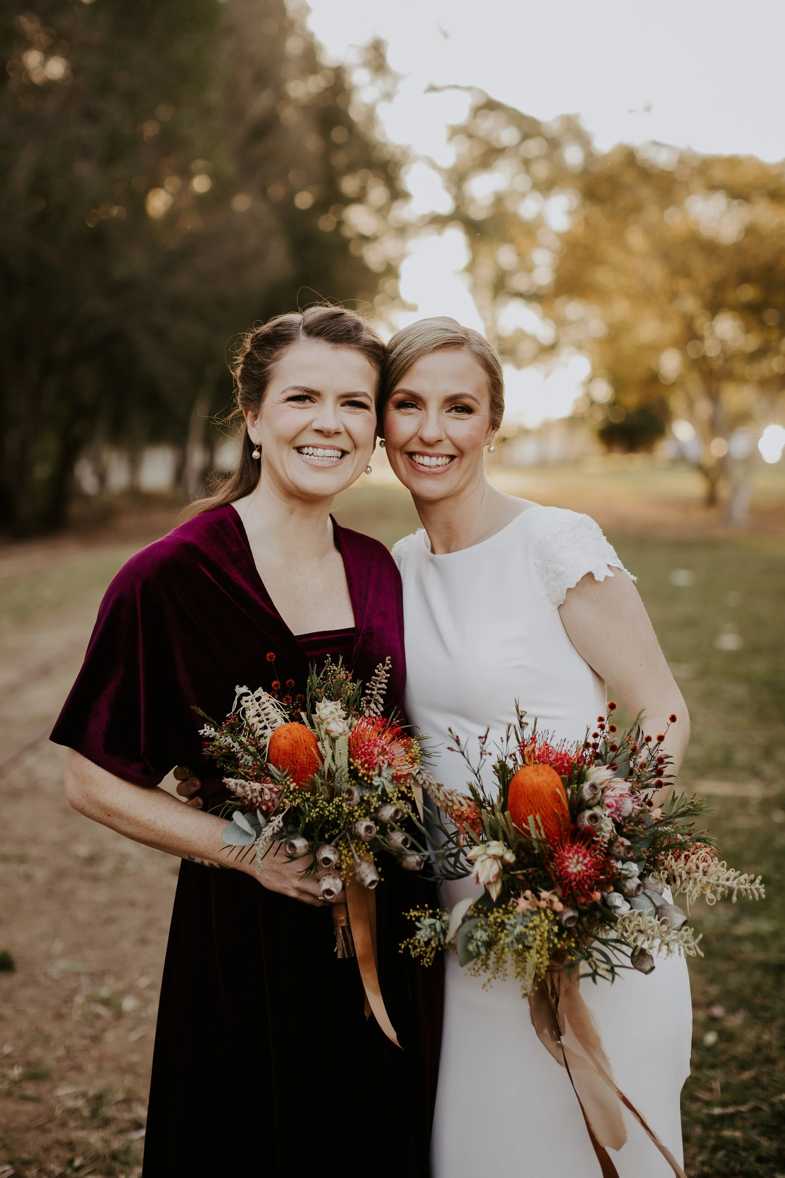 Two women in wedding and formal attire holding bouquets, smiling outdoors with trees and soft sunlight in the background.