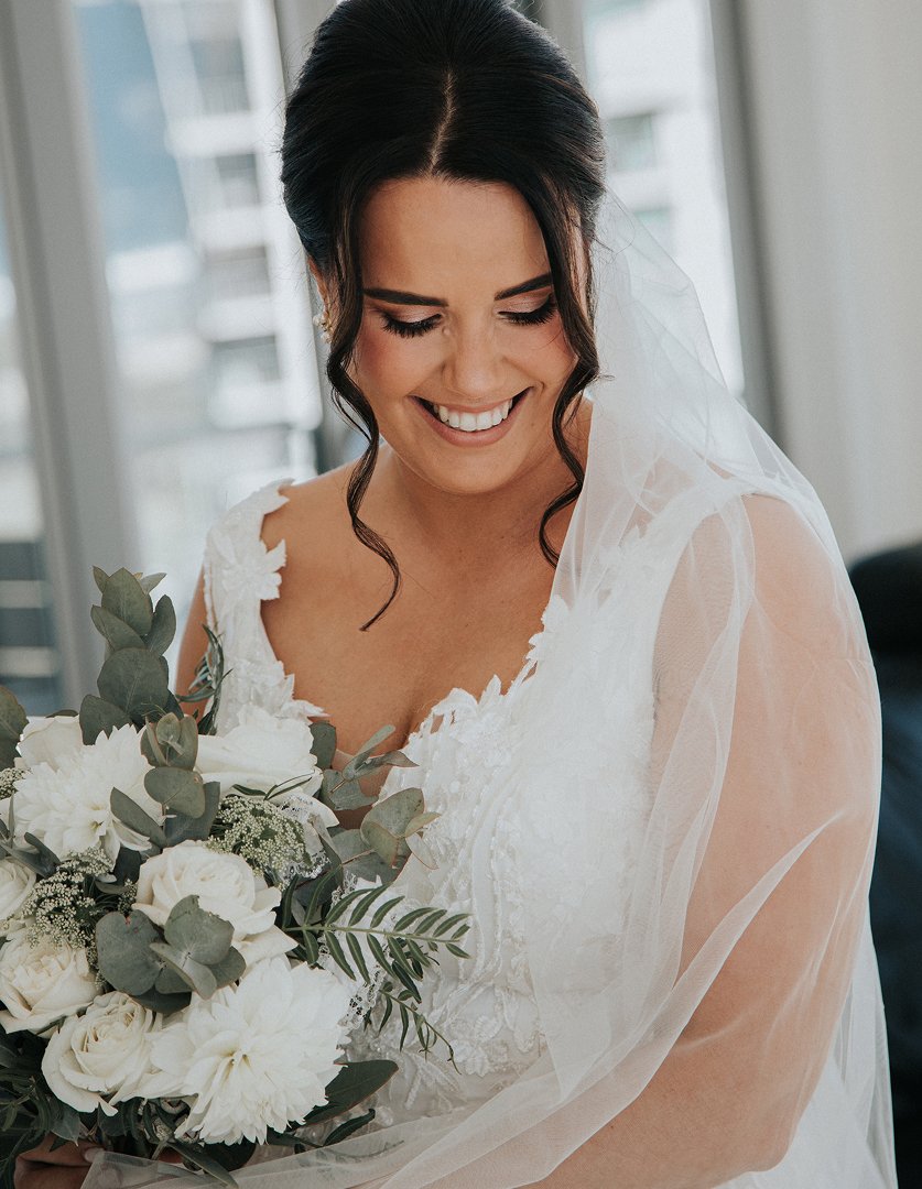 A bride with dark hair styled in loose curls, wearing a white lace wedding dress and veil, smiling while holding a bouquet of white flowers and green foliage.