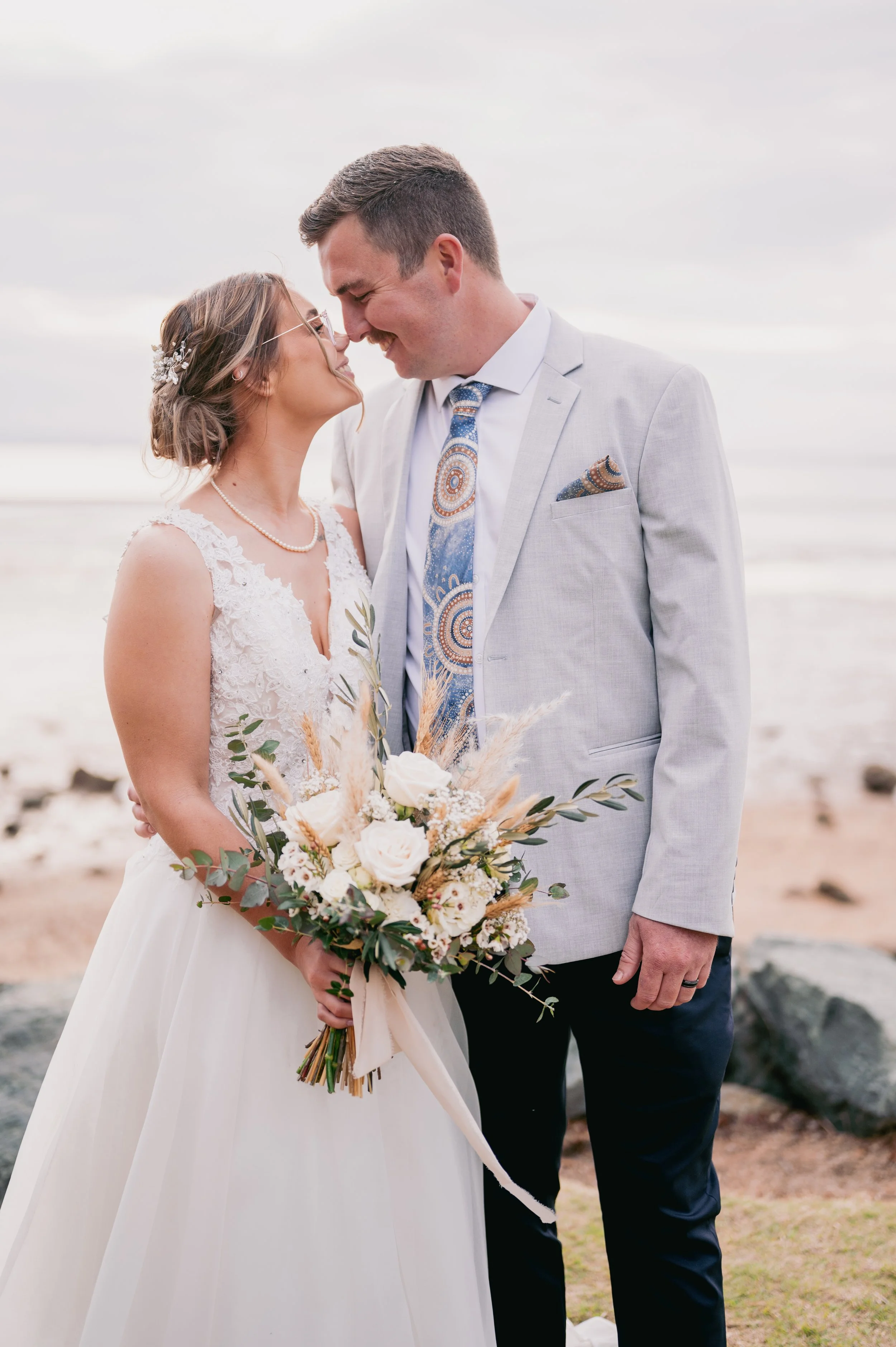 A bride and groom on the beach, leaning their foreheads together, holding a bouquet of white and beige flowers and greenery, dressed in wedding attire, with a cloudy sky background.