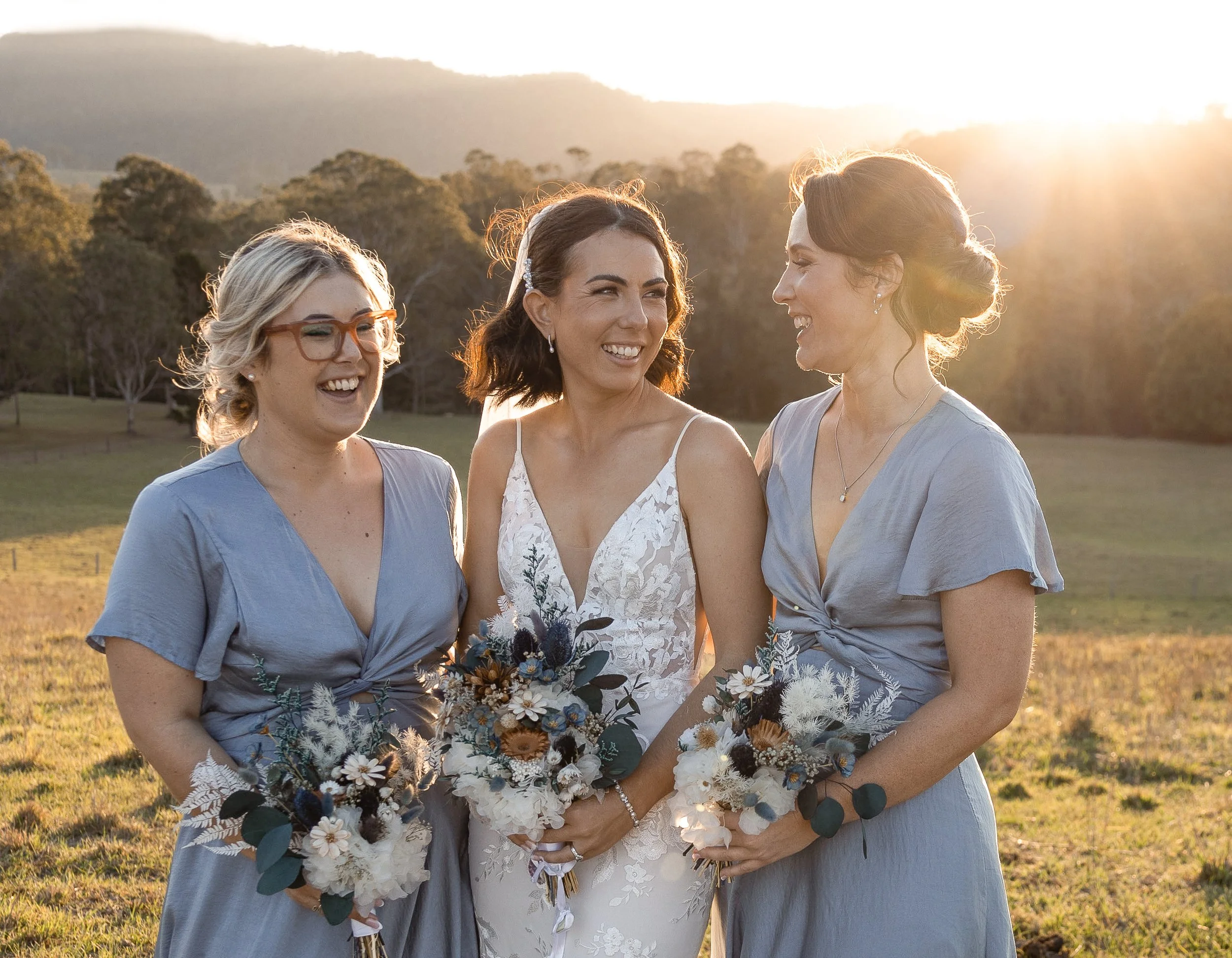 Three women in a field at sunset, with the bride in a white lace dress holding a bouquet, and her two bridesmaids in blue dresses holding smaller bouquets, all smiling and laughing.