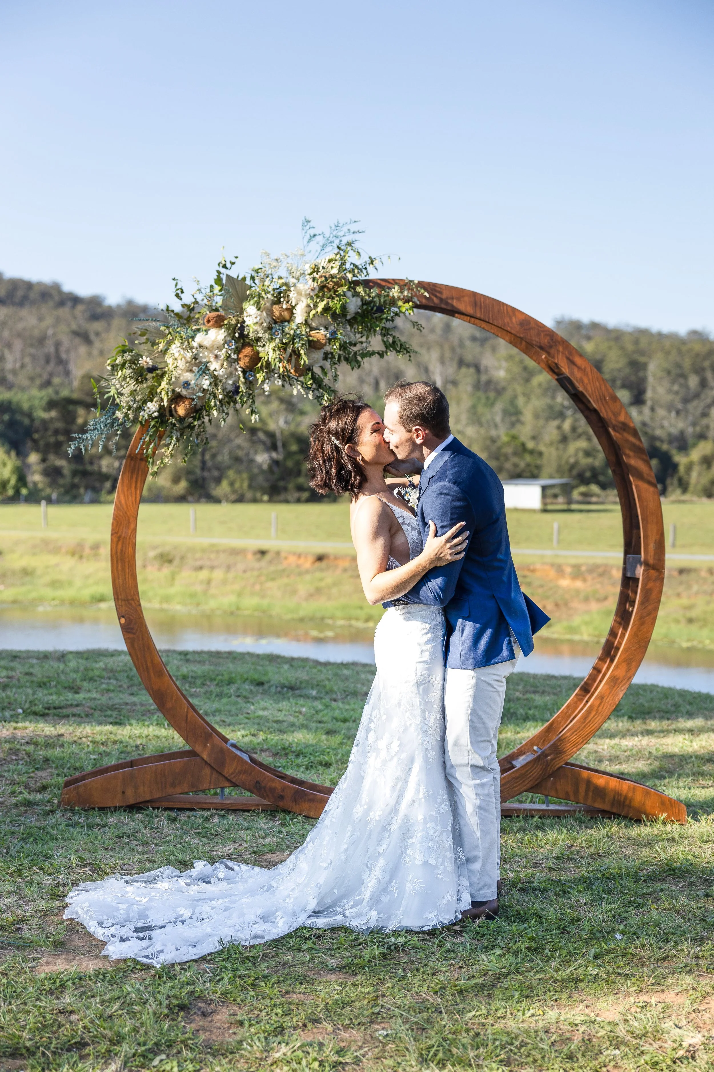 A couple in wedding attire kissing beneath a circular wooden arch decorated with flowers, outdoors on a grassy area with a small body of water and trees in the background.