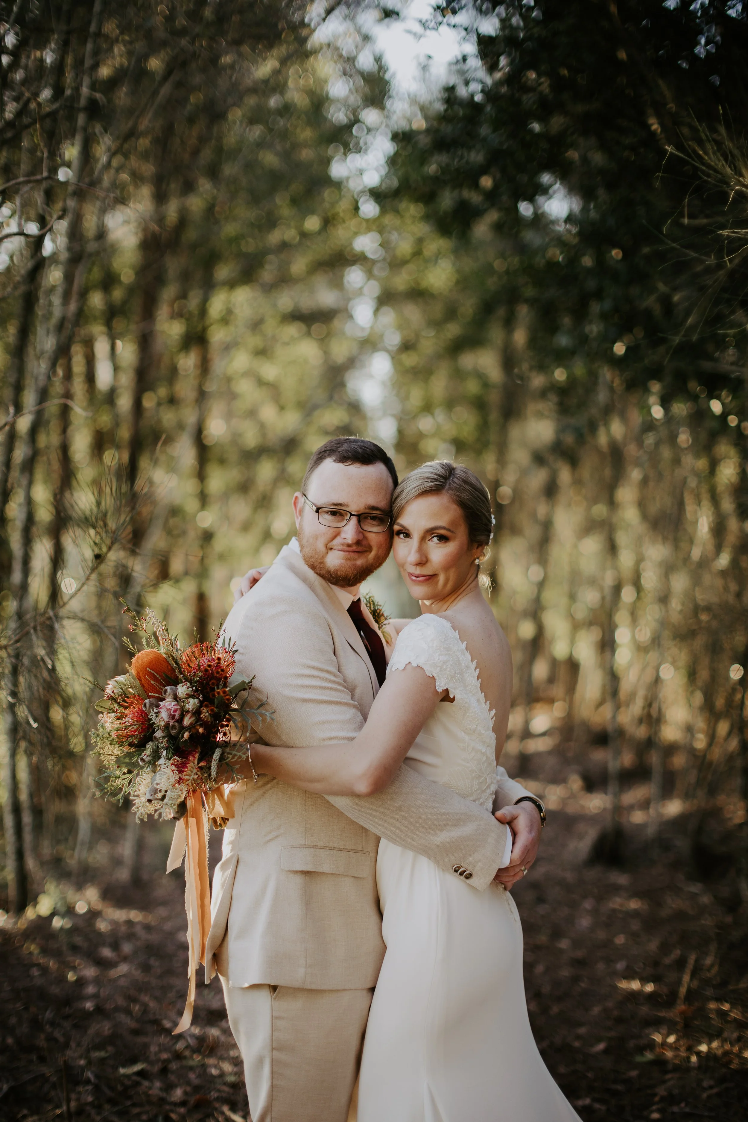 A newlywed couple hugging in a wooded outdoor setting, the bride holding a bouquet of colorful flowers, both dressed in wedding attire, looking at the camera with smiles.