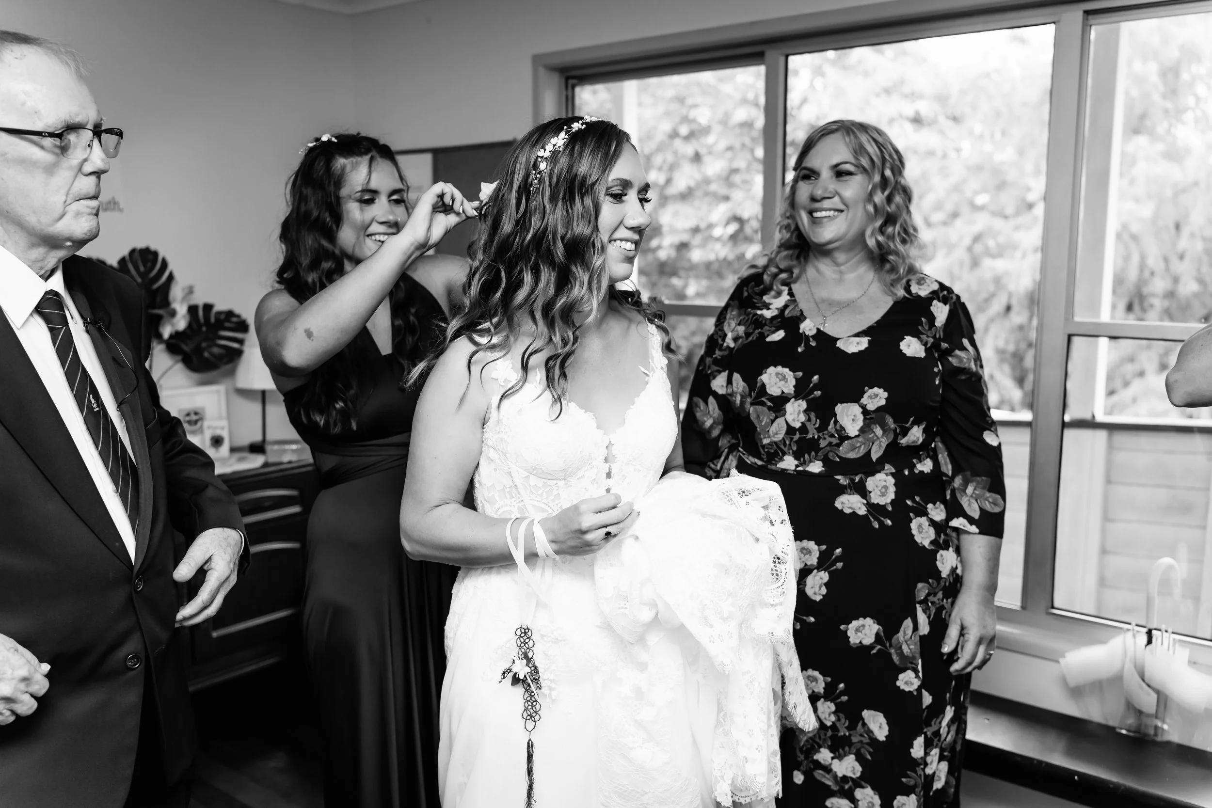 A bride in a white dress holding a lace veil, smiling in a room with large windows, surrounded by women and a man, as she is being helped with her hair and accessories.