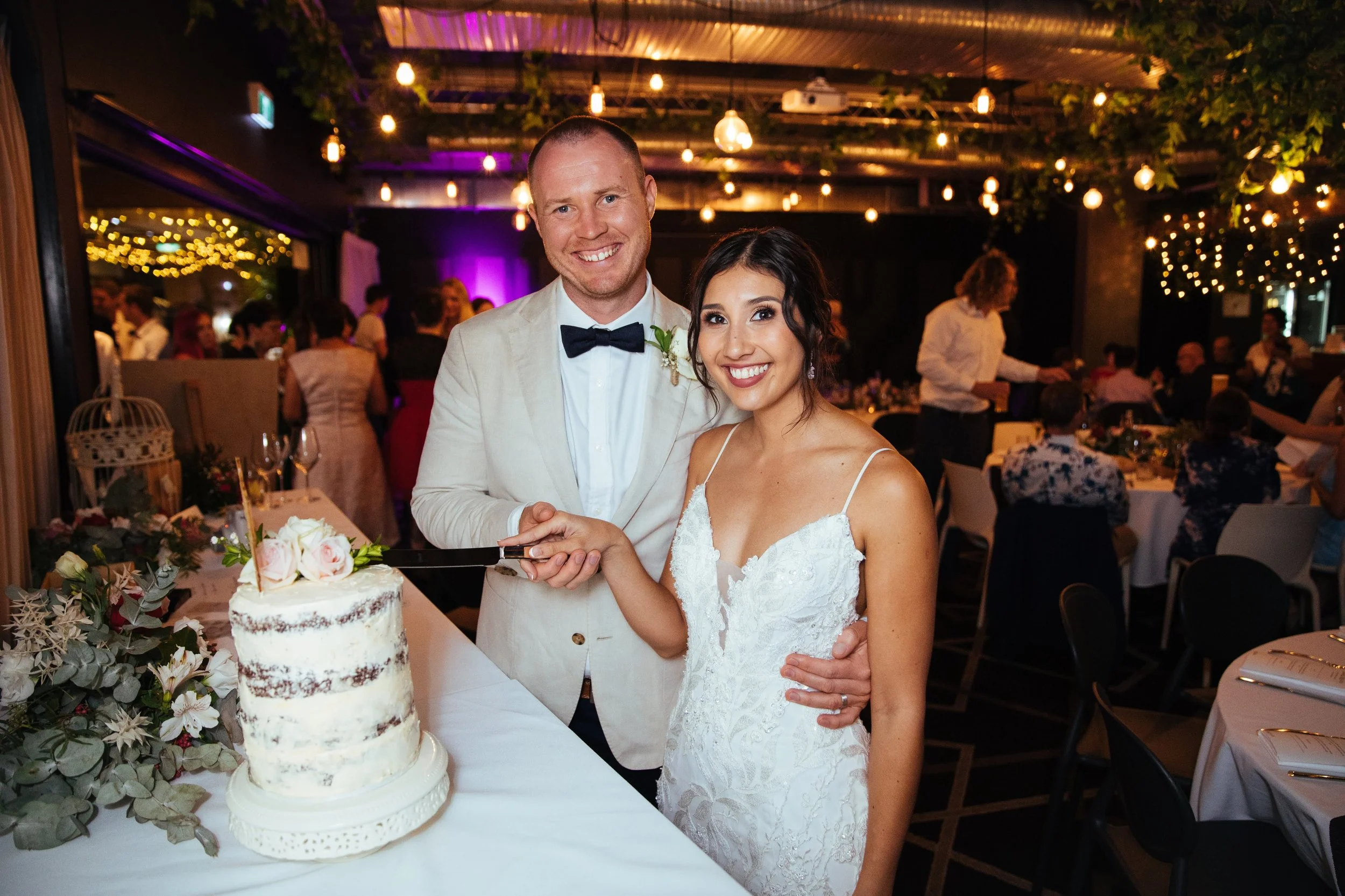A newlywed couple in wedding attire smiling as they cut their wedding cake at a celebration with decorated tables and guests in the background.