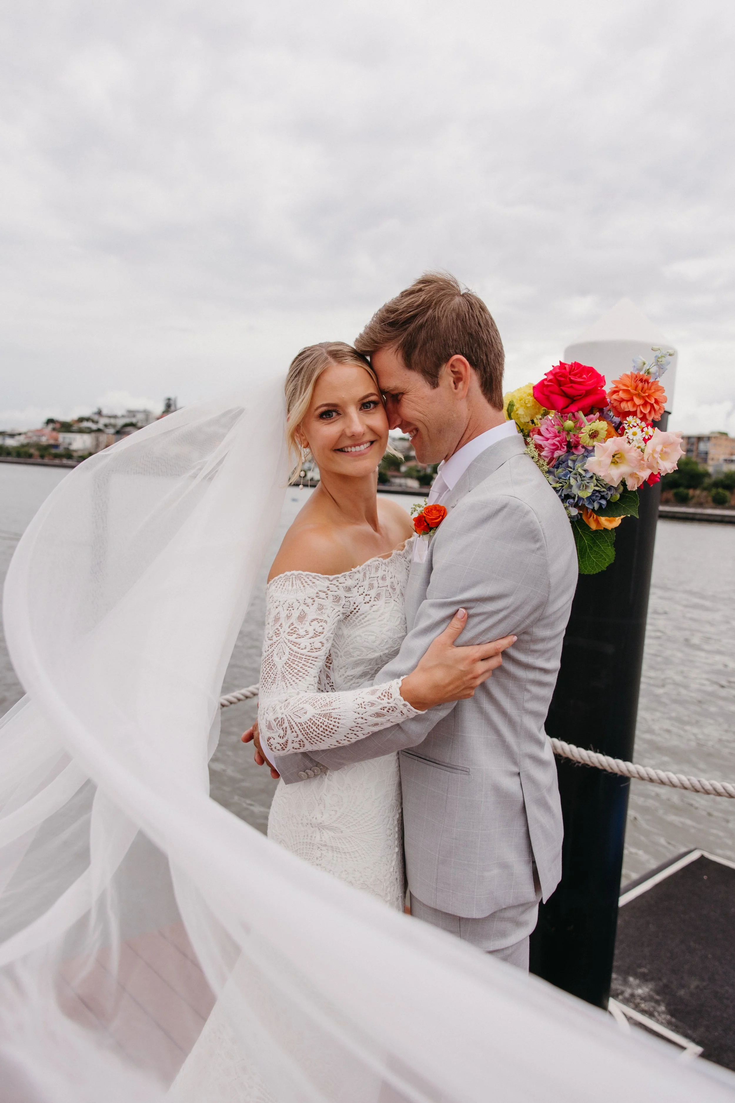 A bride and groom embrace on a waterfront dock, with the bride in a white lace off-shoulder dress and veil, and the groom in a light gray suit, overlooking a river and cityscape.
