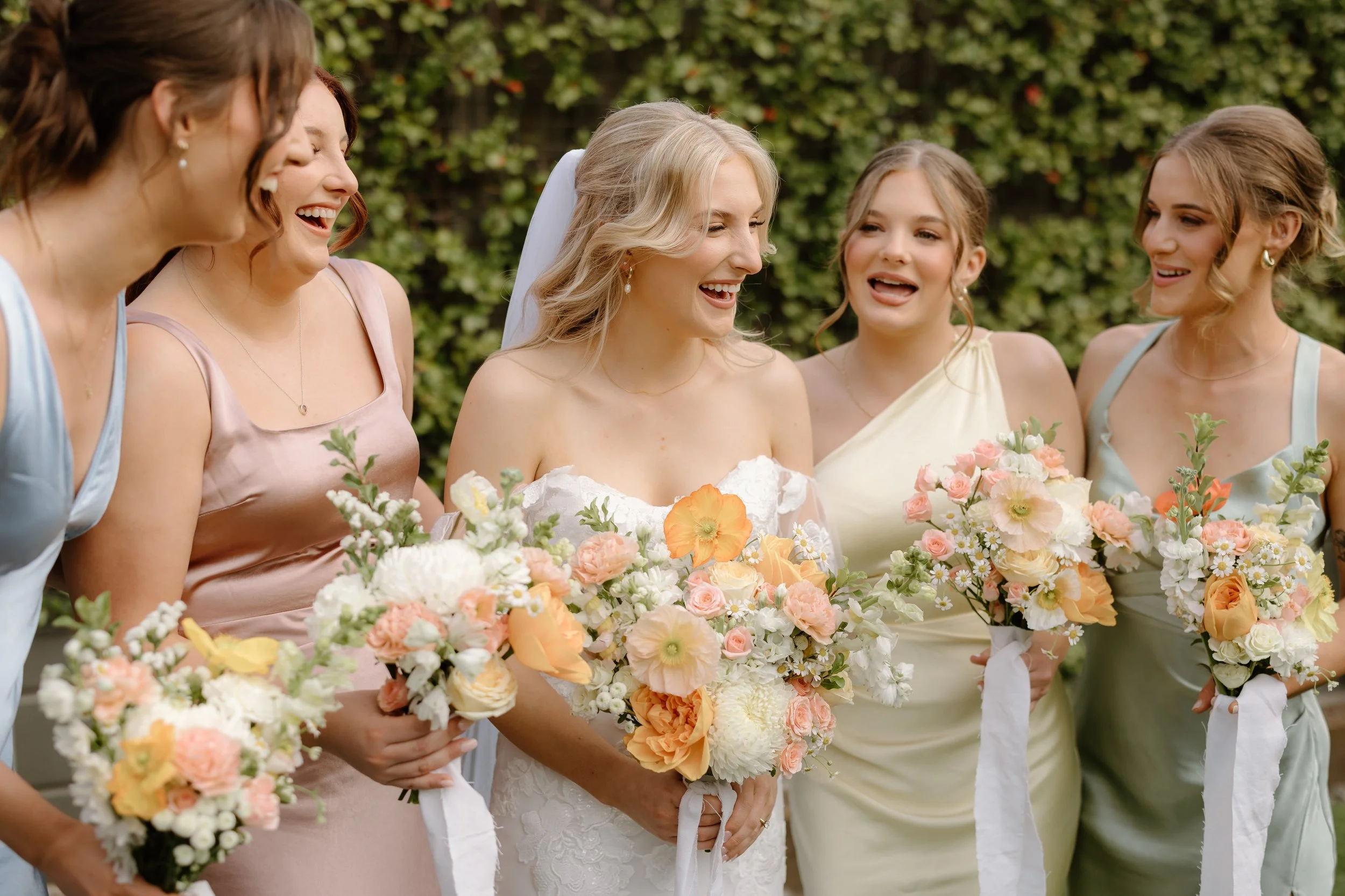 Bride and bridesmaids are smiling and holding bouquets outdoors at a wedding.