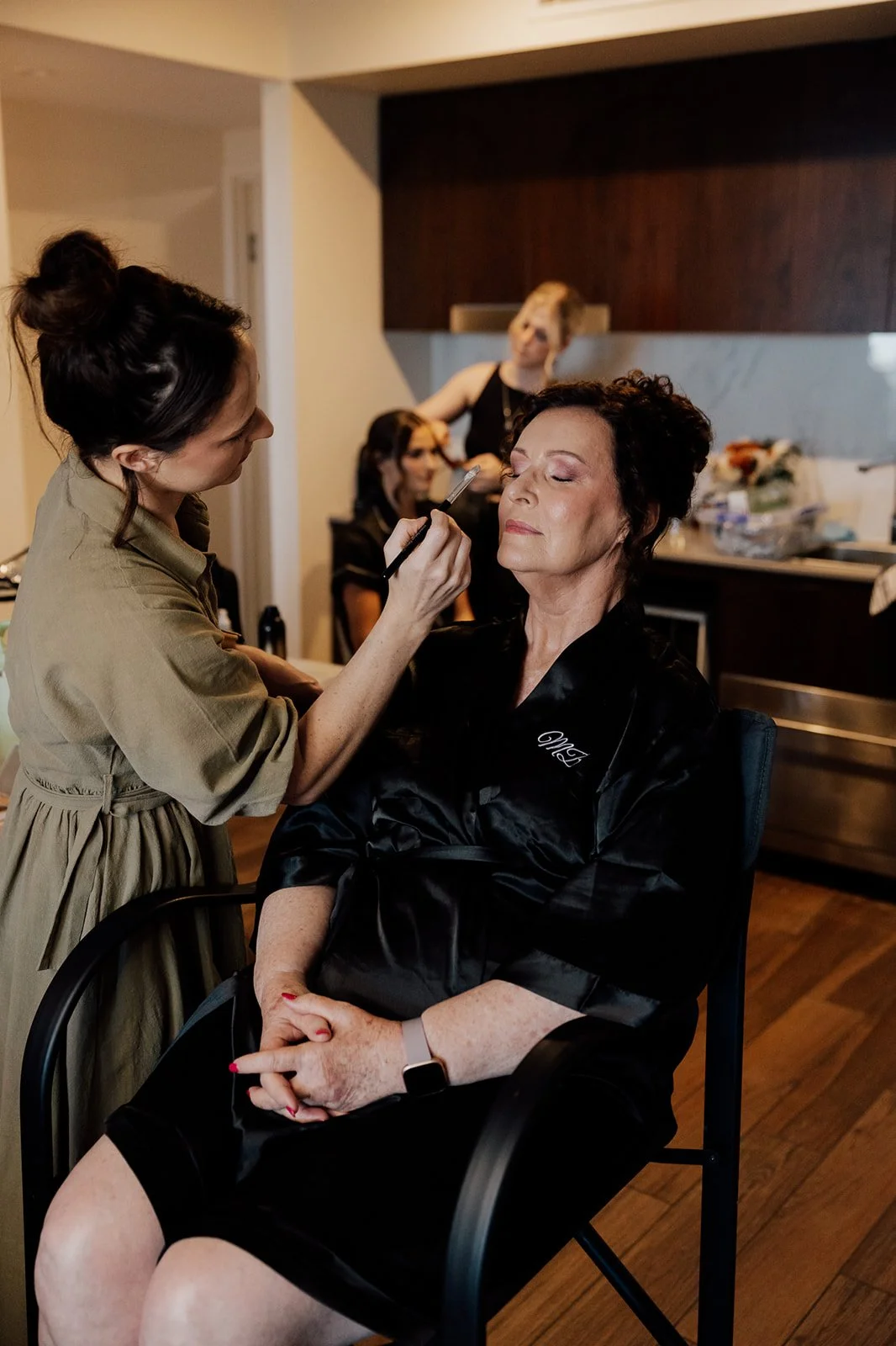 A woman getting her makeup done by a makeup artist in a room with other women, someone in the background is working on their makeup.