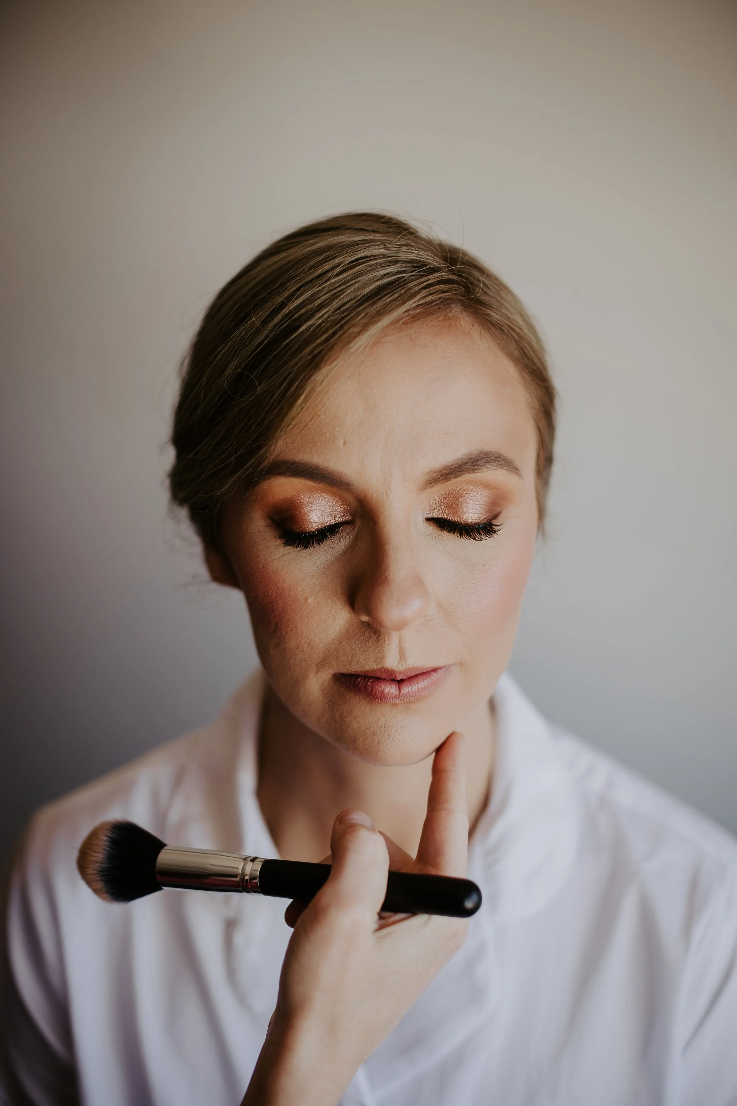 A woman with makeup and short hair, wearing a white shirt, has her eyes closed while a makeup brush is gently touching her chin.