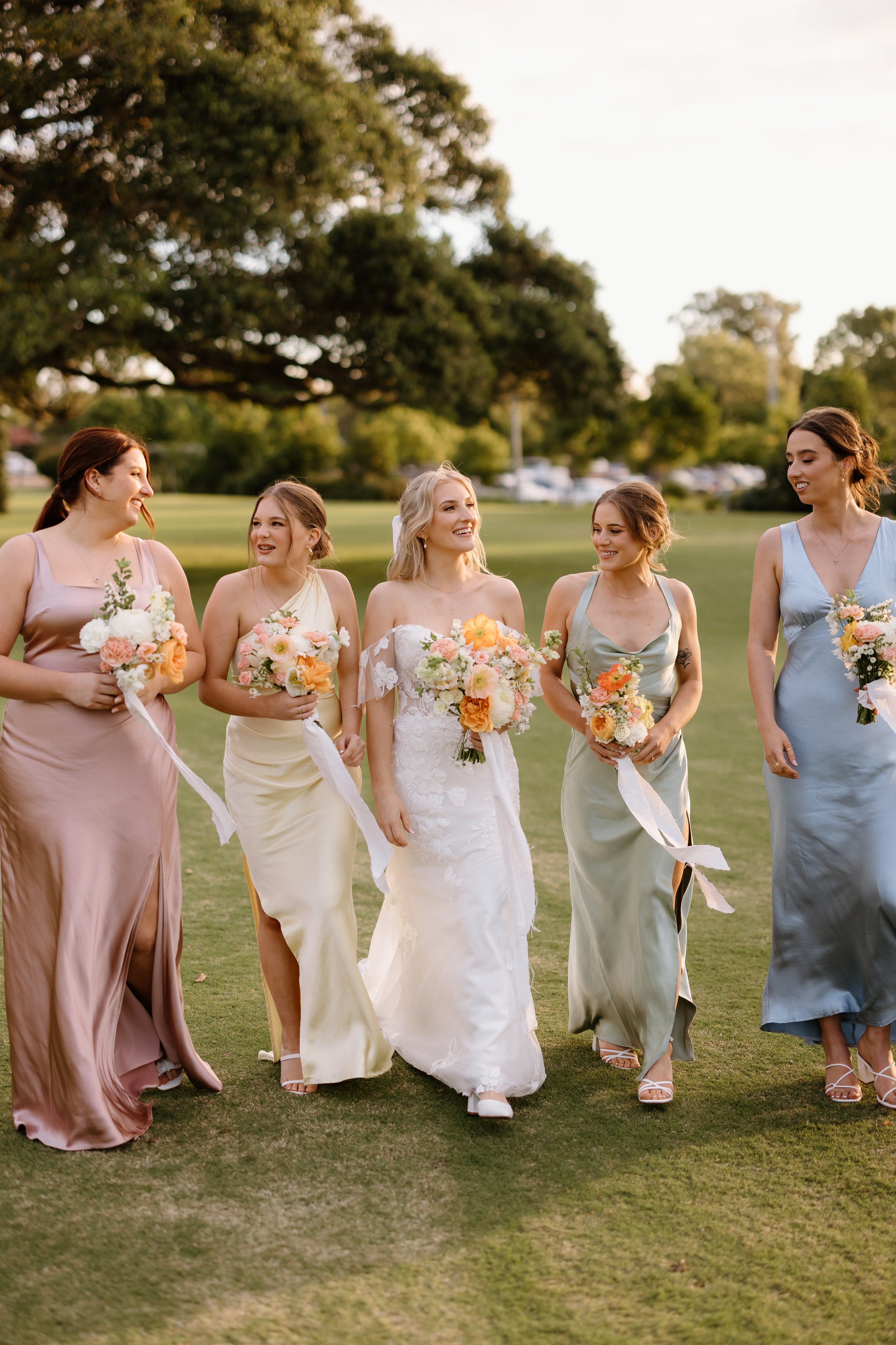 A bride and five bridesmaids walking on a grassy outdoor area, holding bouquets of flowers, during a wedding celebration at sunset.
