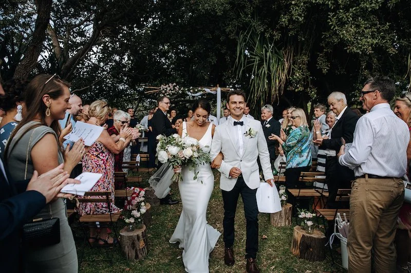 Bride and groom walking down the aisle at an outdoor wedding ceremony, surrounded by seated guests applauding and smiling, with trees and greenery in the background.