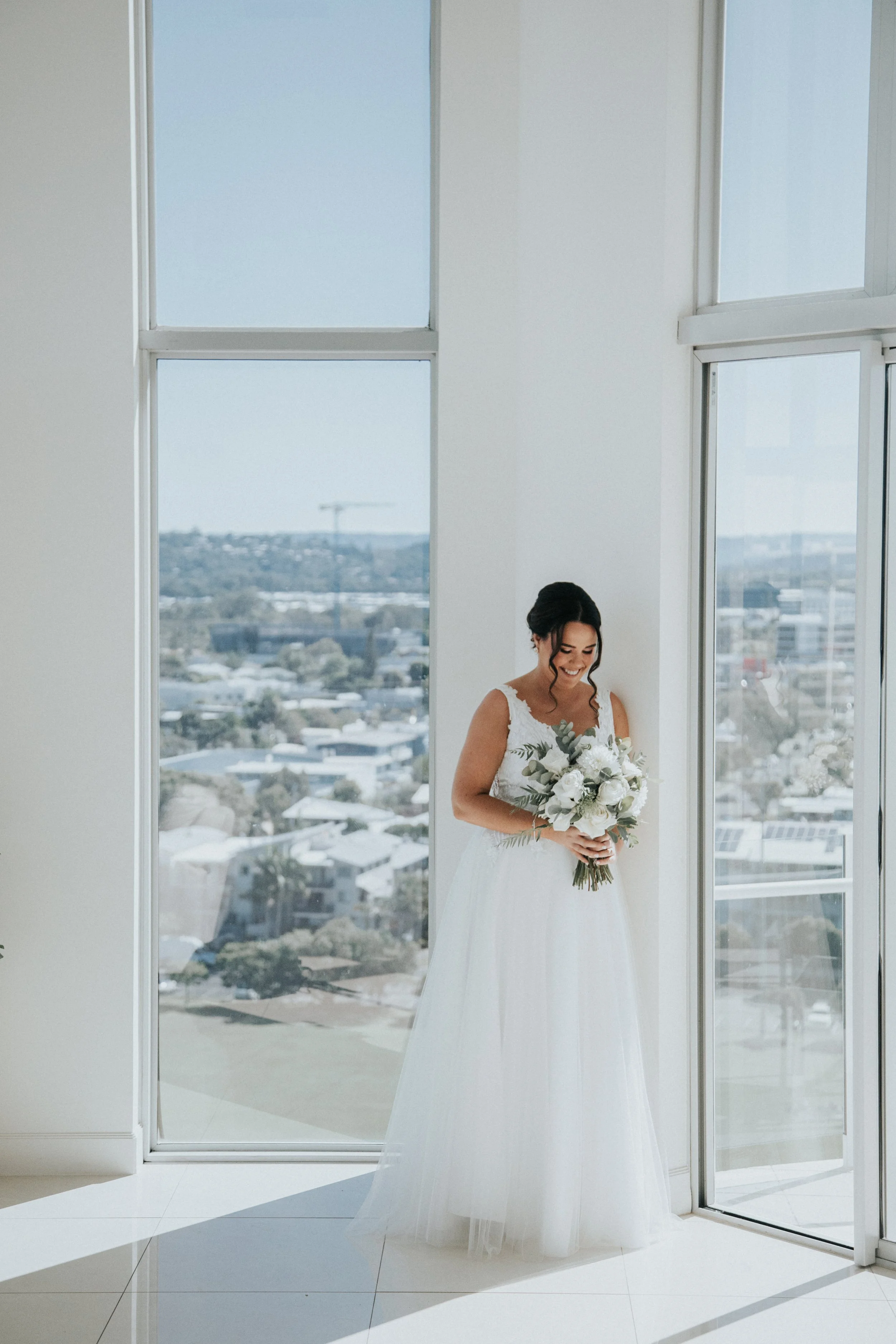 A bride in a white wedding dress holding a bouquet of white flowers, standing by large windows with a city view.