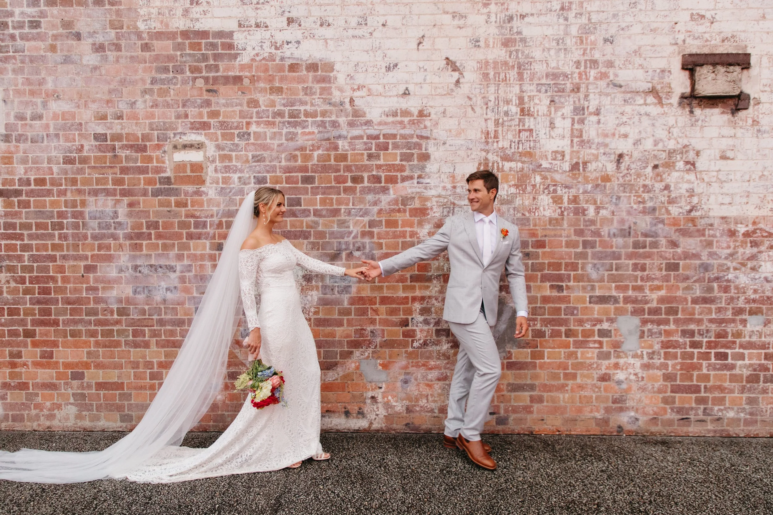 A bride and groom holding hands and smiling against a brick wall. The bride is in a white lace wedding dress with a long train and veil, holding a bouquet of flowers. The groom is in a light gray suit with brown shoes.